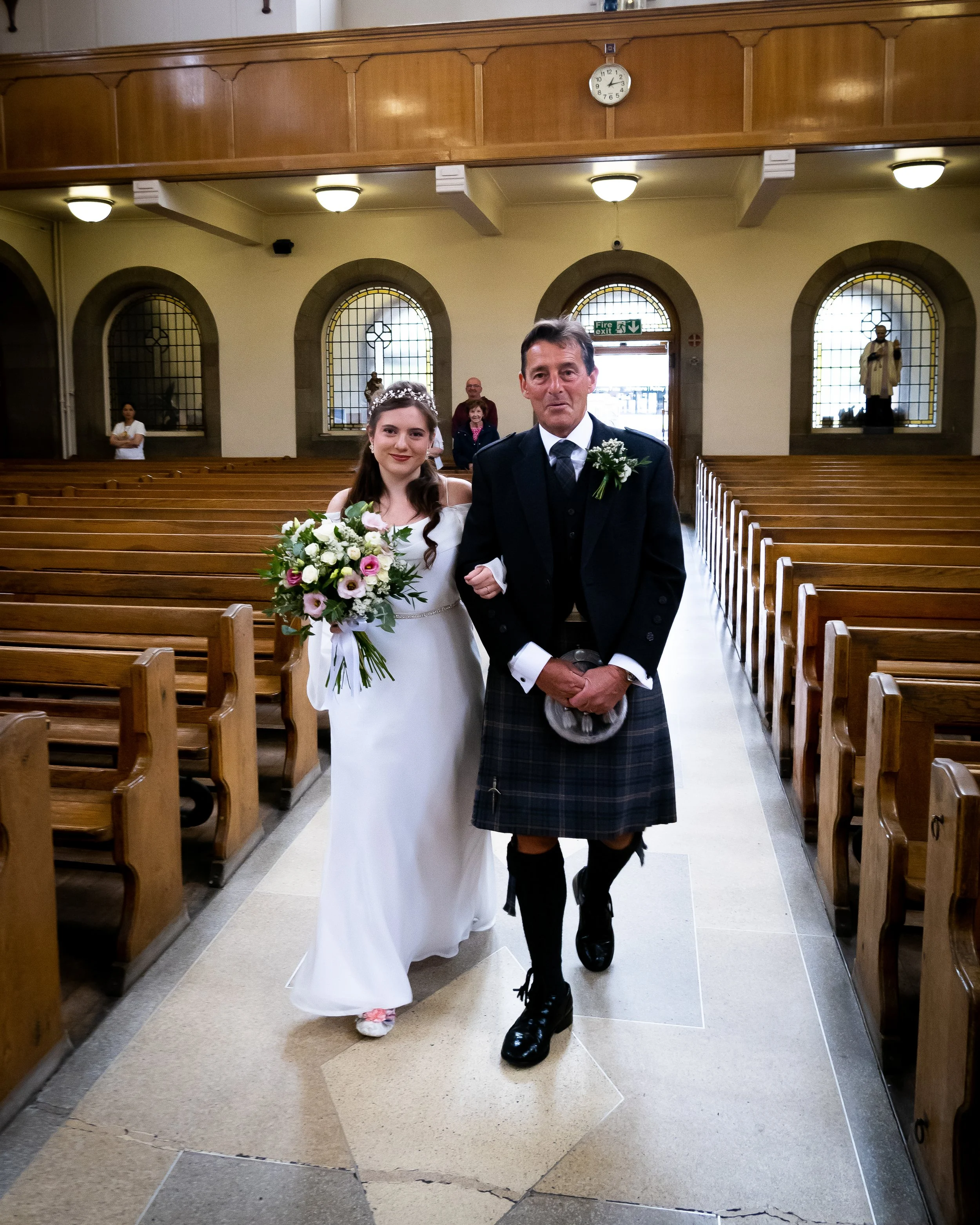 A bride in a white wedding dress holding a bouquet of flowers walking arm-in-arm with a man in traditional Scottish attire, inside a church with wooden pews and stained glass windows.