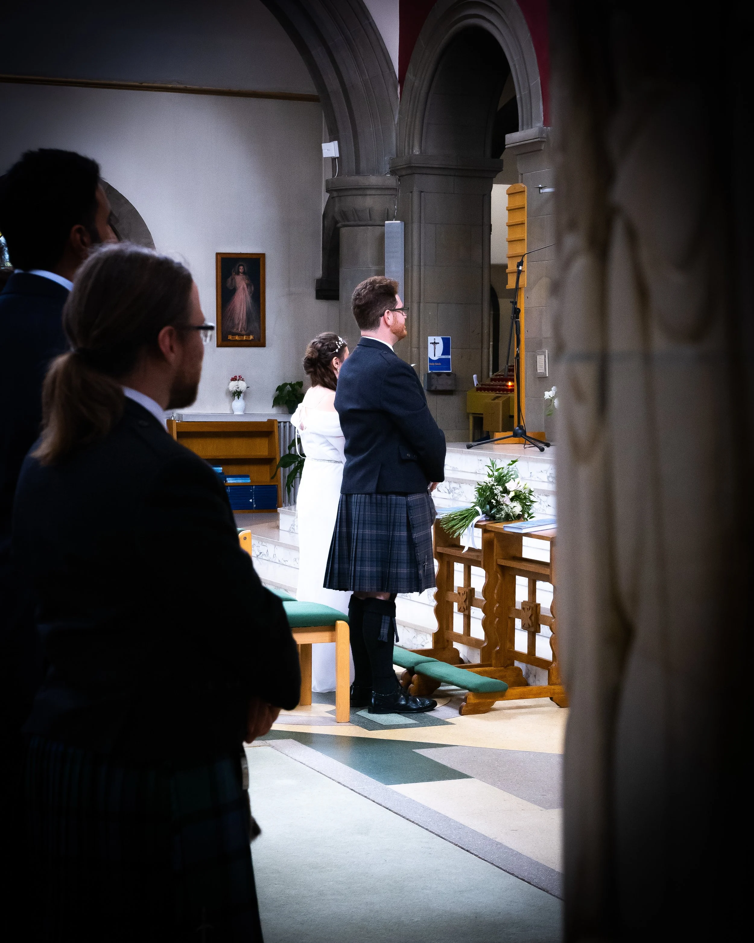 People attending a church service, with one man in a kilt standing near the altar, others seated and praying, inside a church with religious artwork and flowers.