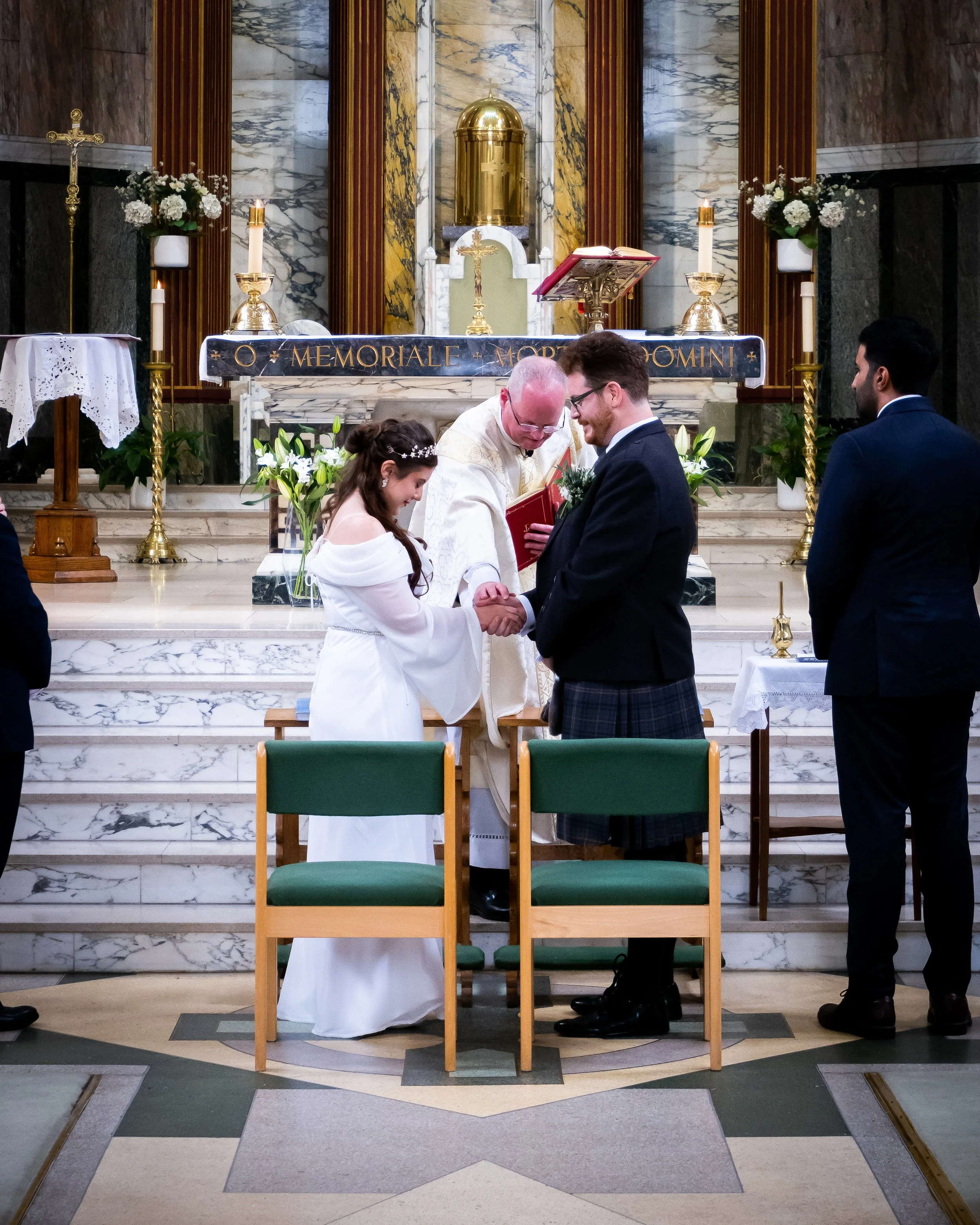 A couple getting married during a church wedding ceremony, with a priest officiating and others present, in a church with marble altar, flowers, and religious symbols.