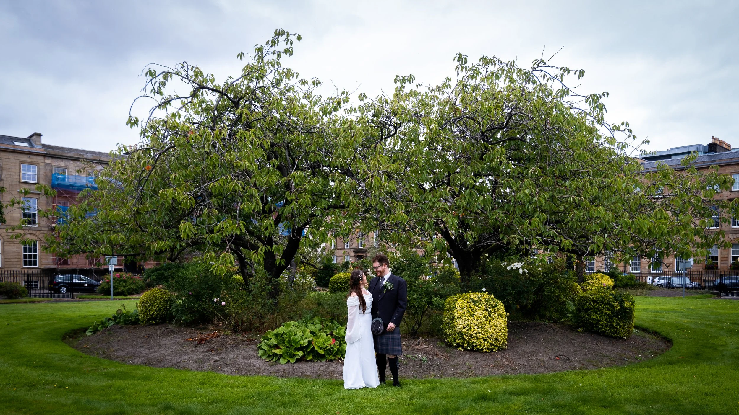 A couple dressed in wedding attire standing in a park with well-manicured grass, bushes, and a large tree in the background. The woman is wearing a white dress and the man is in a traditional Scottish kilt with a black jacket. They are looking at eac