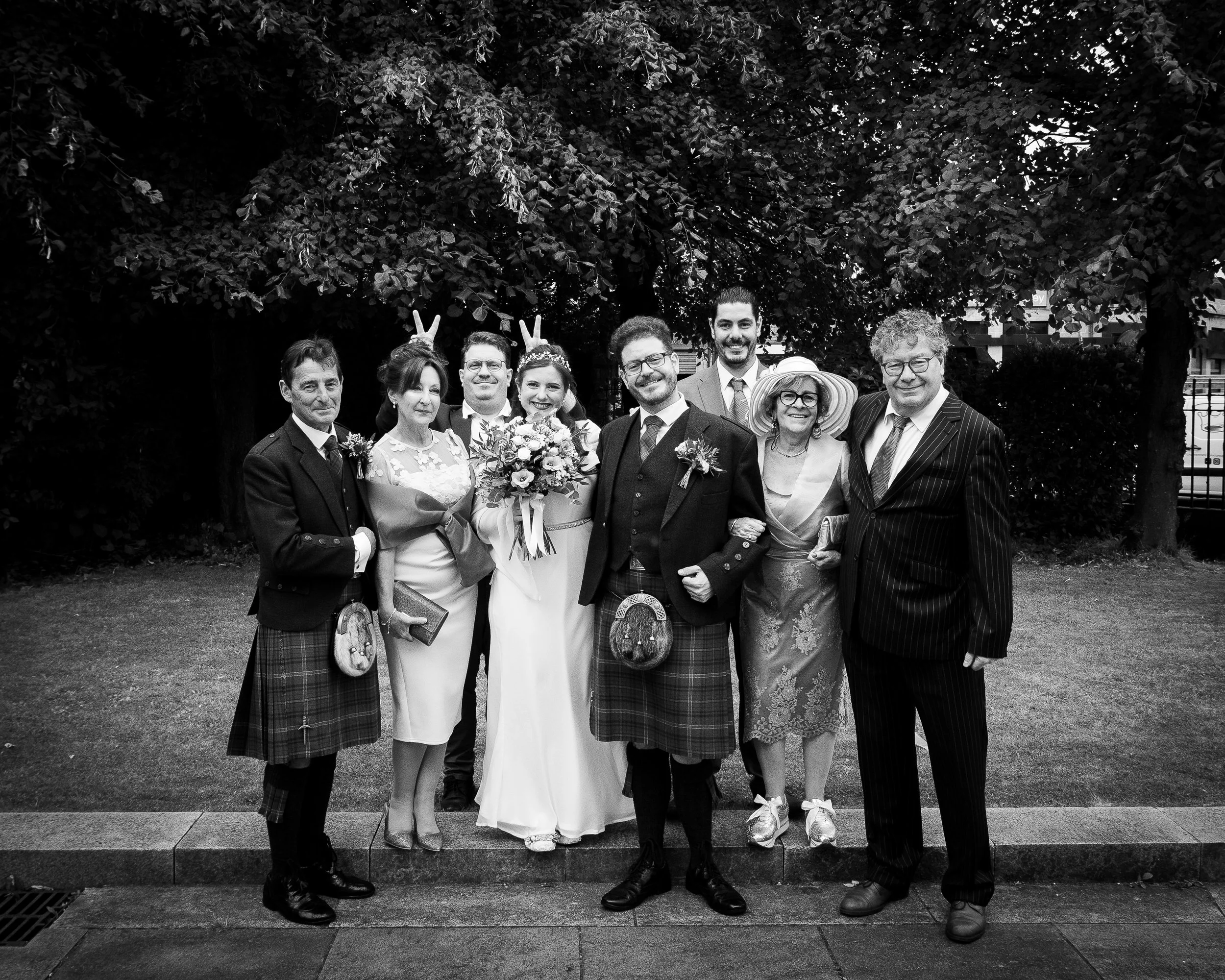 A group of nine people dressed in formal and traditional Scottish attire posing outdoors in front of trees, with some making playful gestures in a black and white photo.