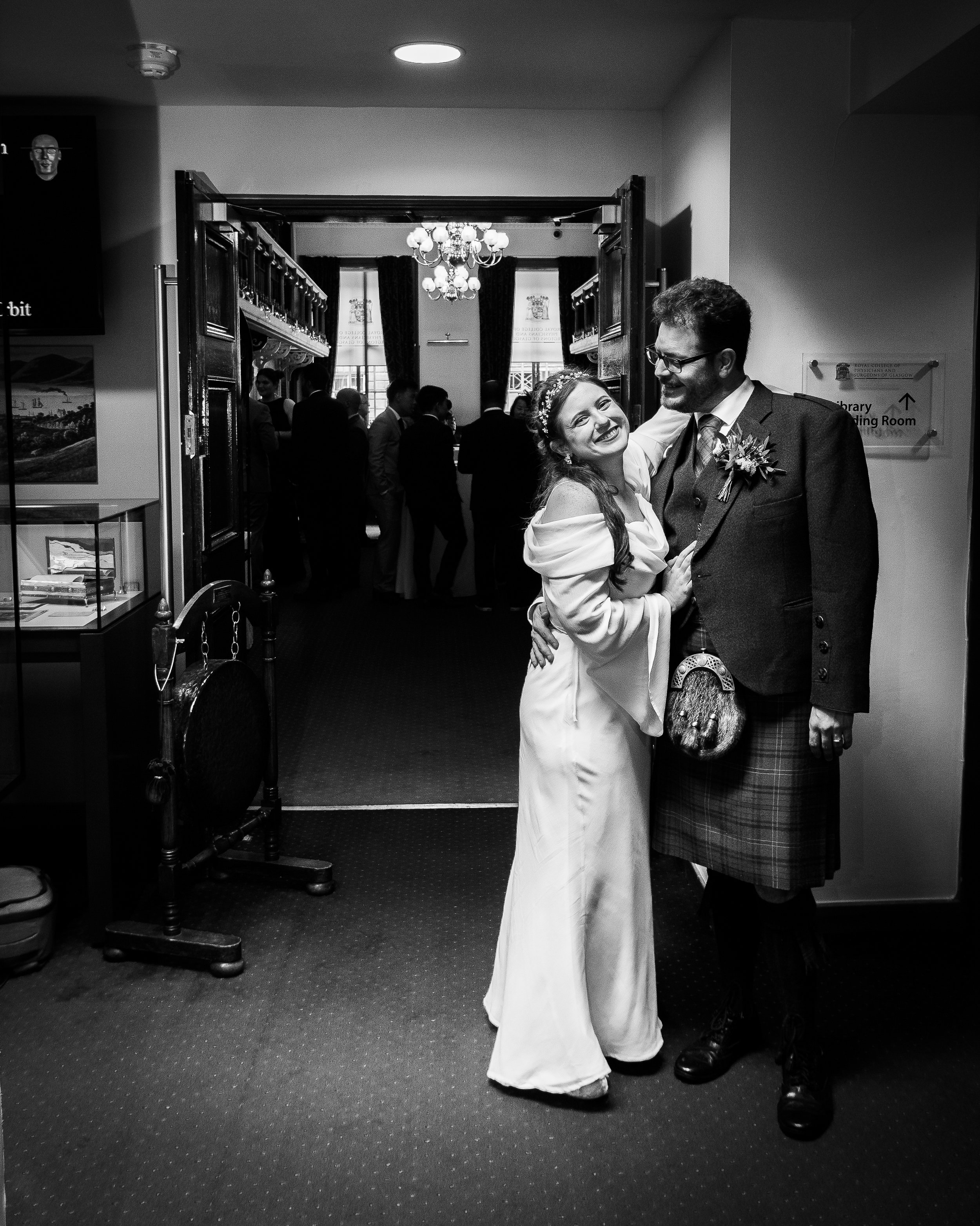 A bride in a white dress and a groom in a kilt are smiling and hugging each other in a hallway at a wedding reception, with guests in the background.