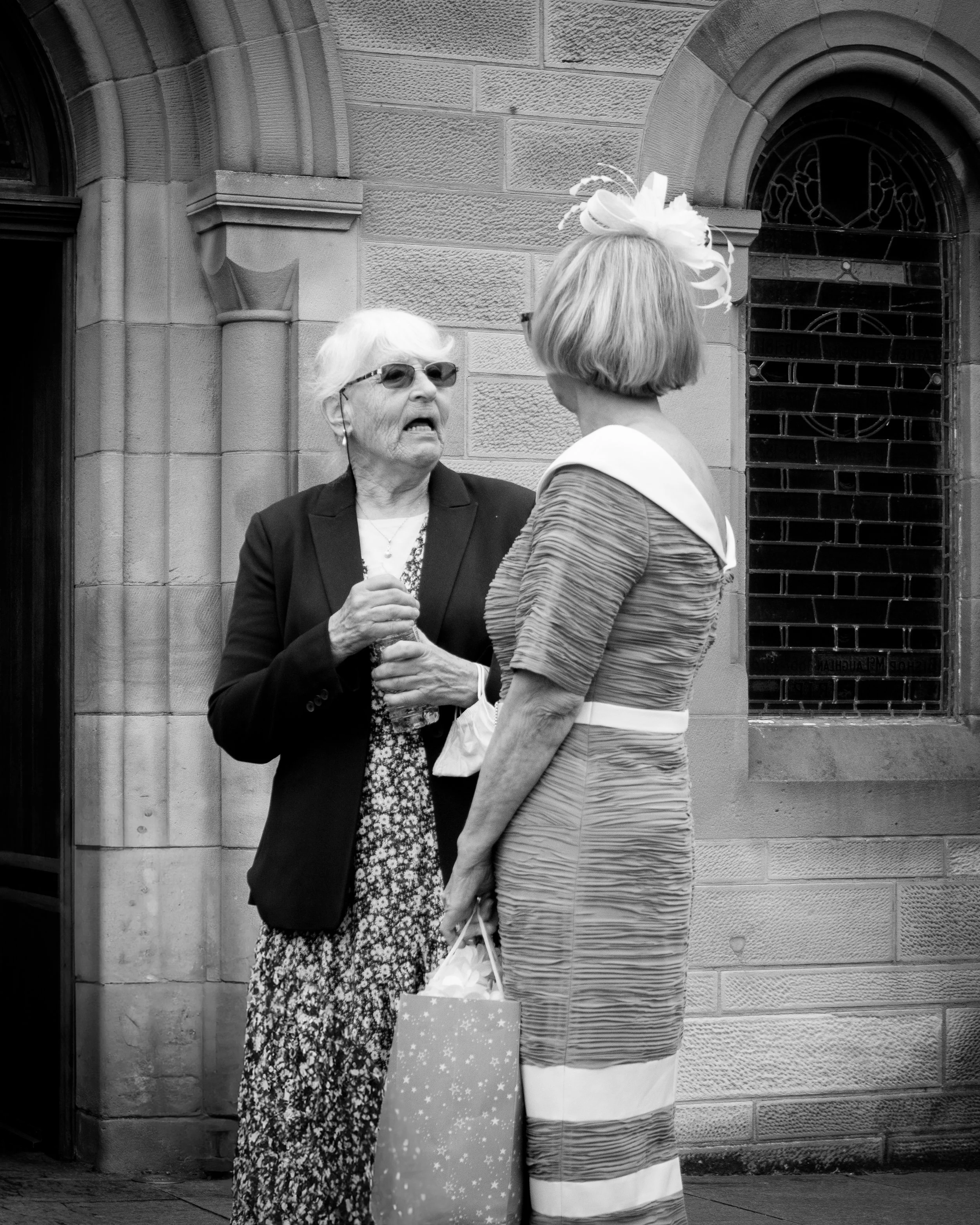 Two women in conversation outside a stone building with arched windows. One is elderly with sunglasses, short white hair, wearing a dark blazer and patterned dress, holding a water bottle. The other has a short bob haircut, wearing a striped dress wi