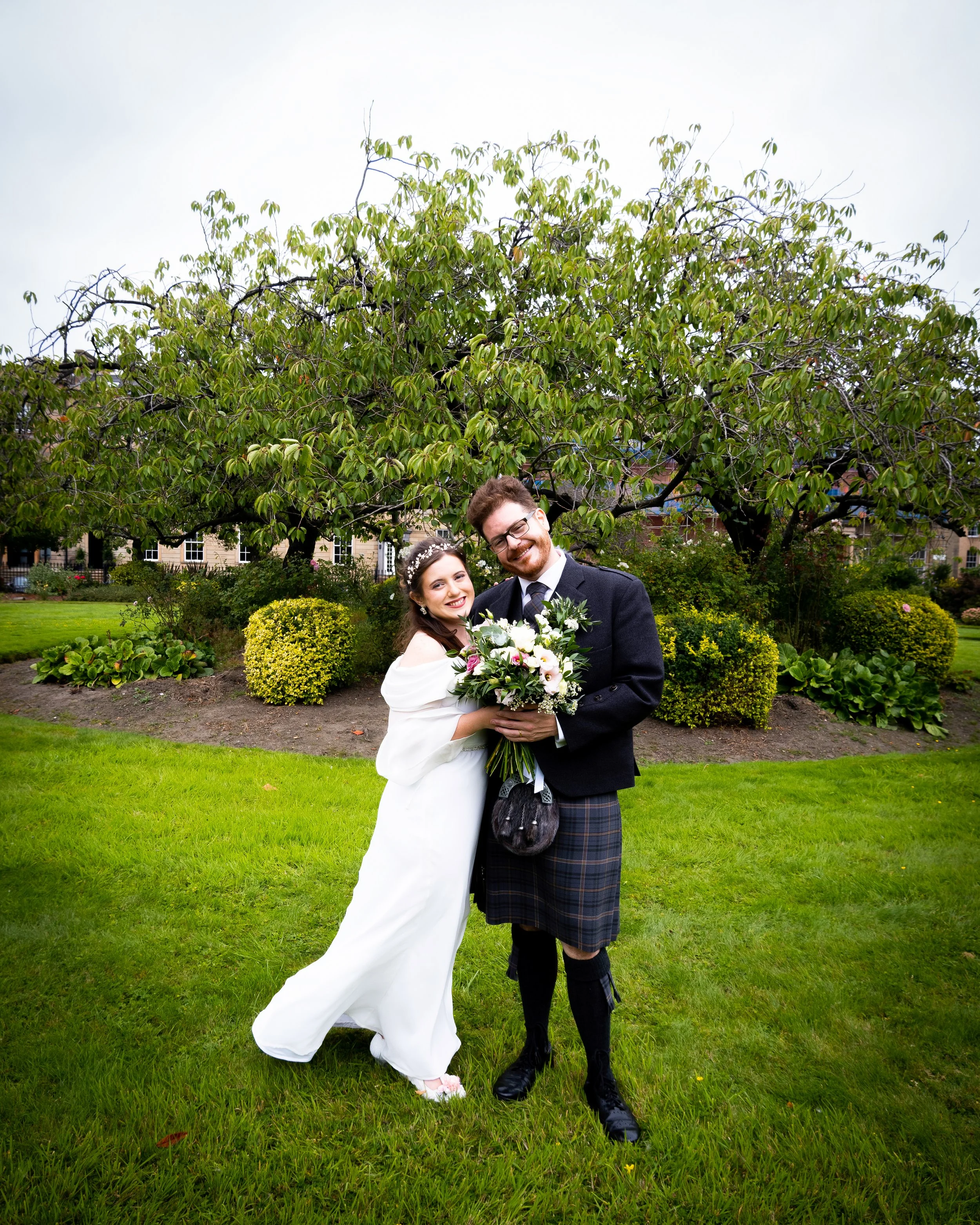 A bride and groom standing on a lush green lawn under a tree, holding a bouquet of flowers, smiling for a wedding photo.