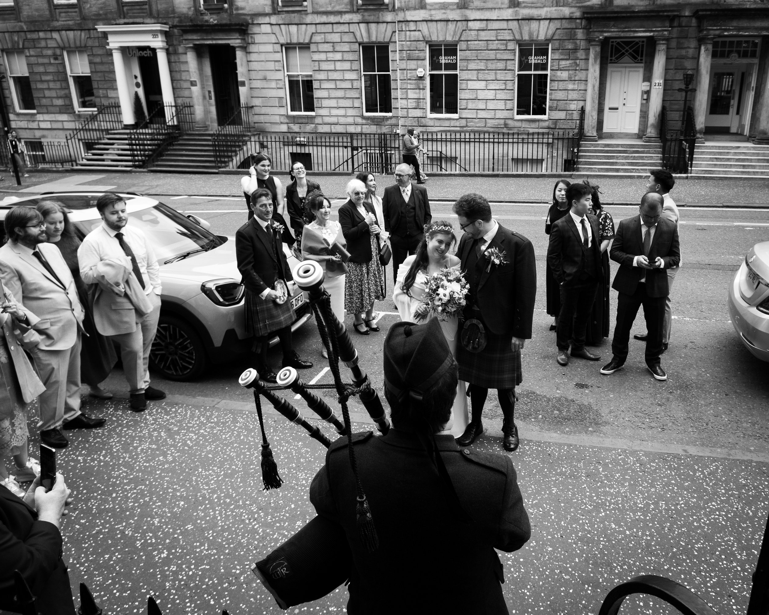 A group of people gather outside a building for a wedding, with the bride and groom in the center, the bride holding a bouquet and smiling, while the groom wears a kilt. Men and women in formal attire stand on the street, some using their phones, wit