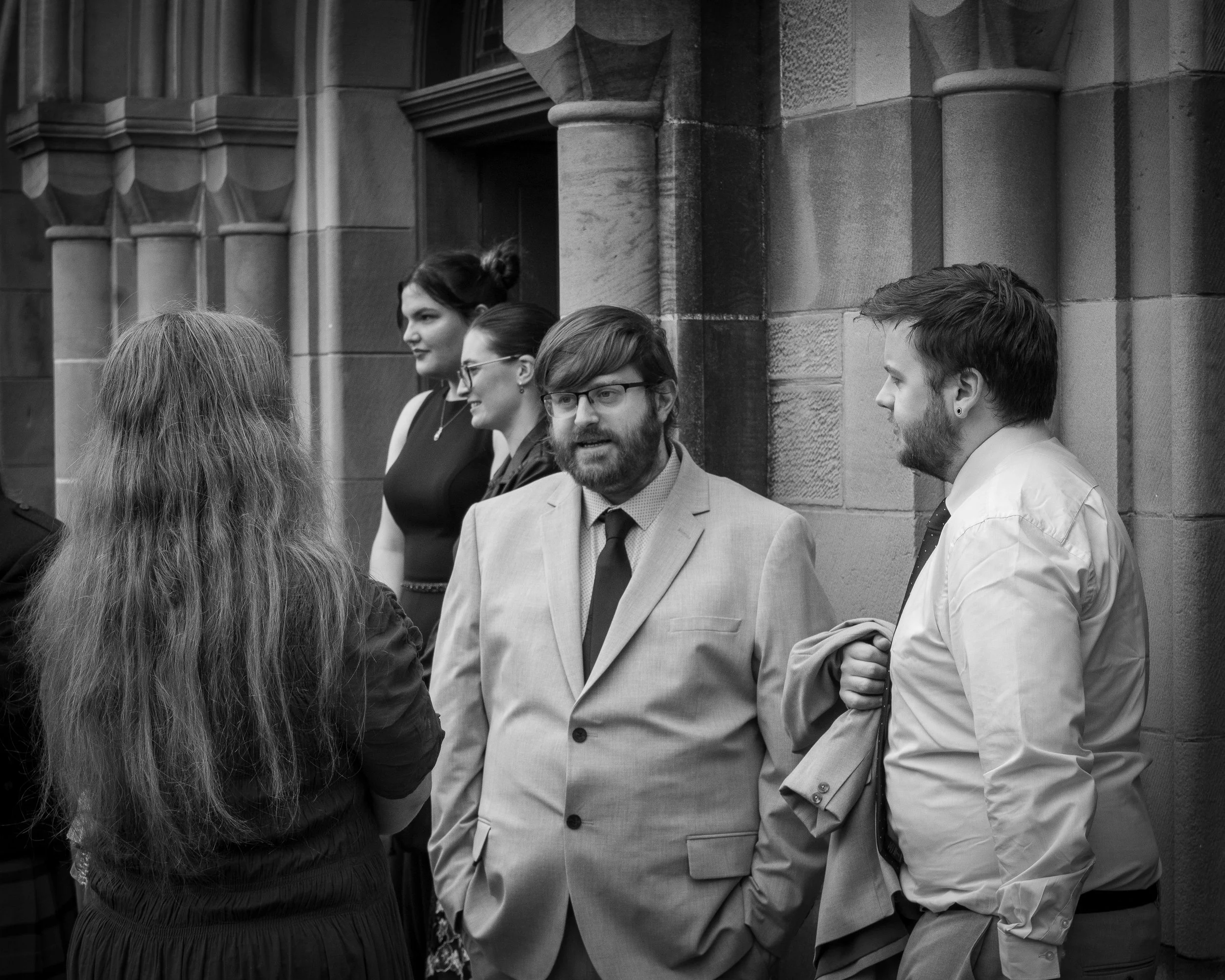 Group of five young adults in formal attire conversing outside a stone building, black-and-white photograph.