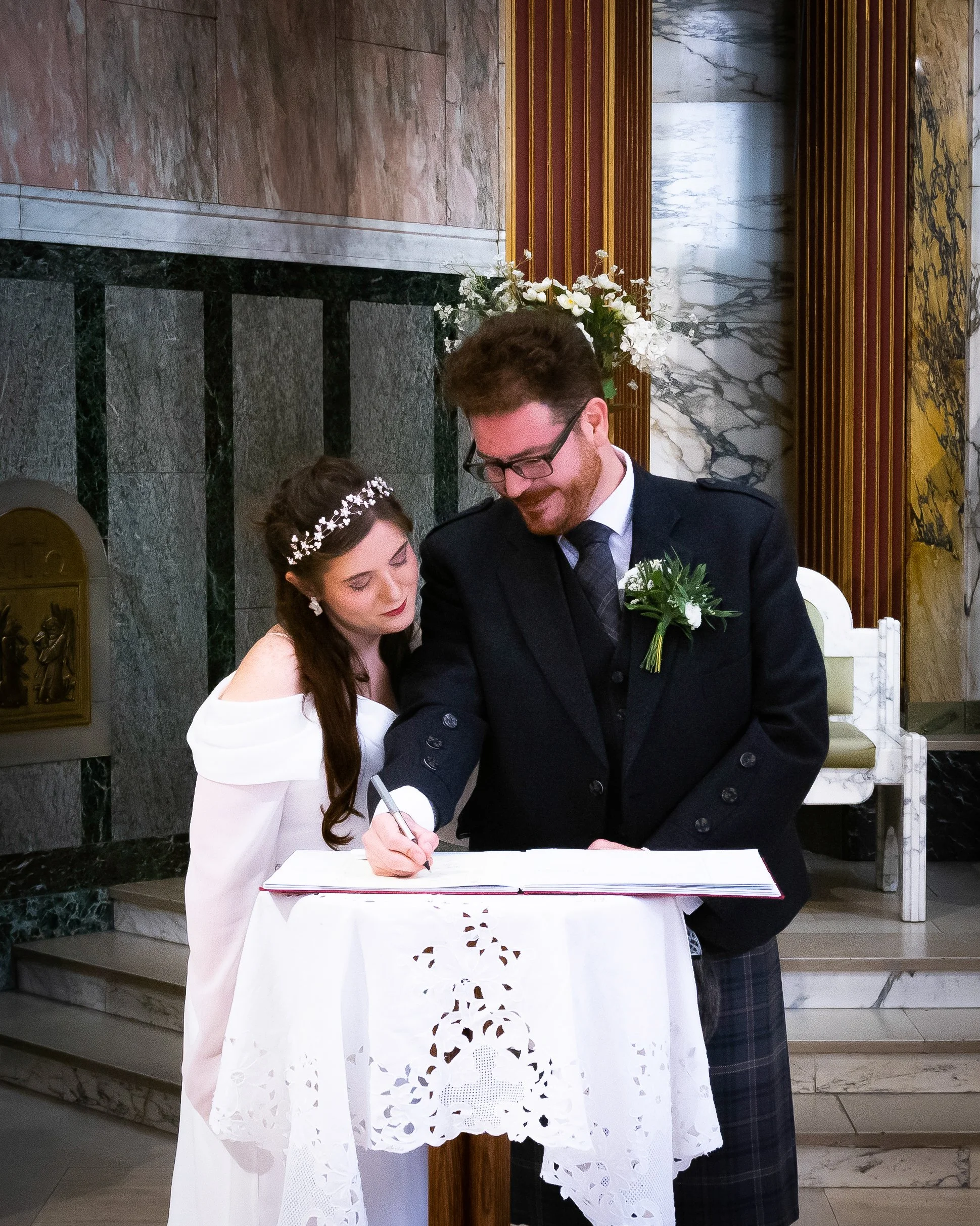 A couple signs a marriage register during their wedding ceremony inside a church. The bride is dressed in a white gown with off-shoulder sleeves, and the groom is wearing a dark suit with a boutonniere. They are leaning over a small table covered wit