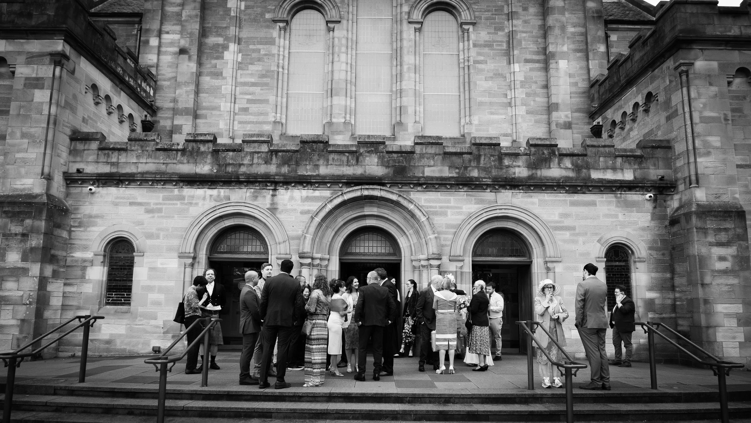 A group of people dressed formally gathering outside a historic stone church or cathedral with arched entryways and large stained glass windows.