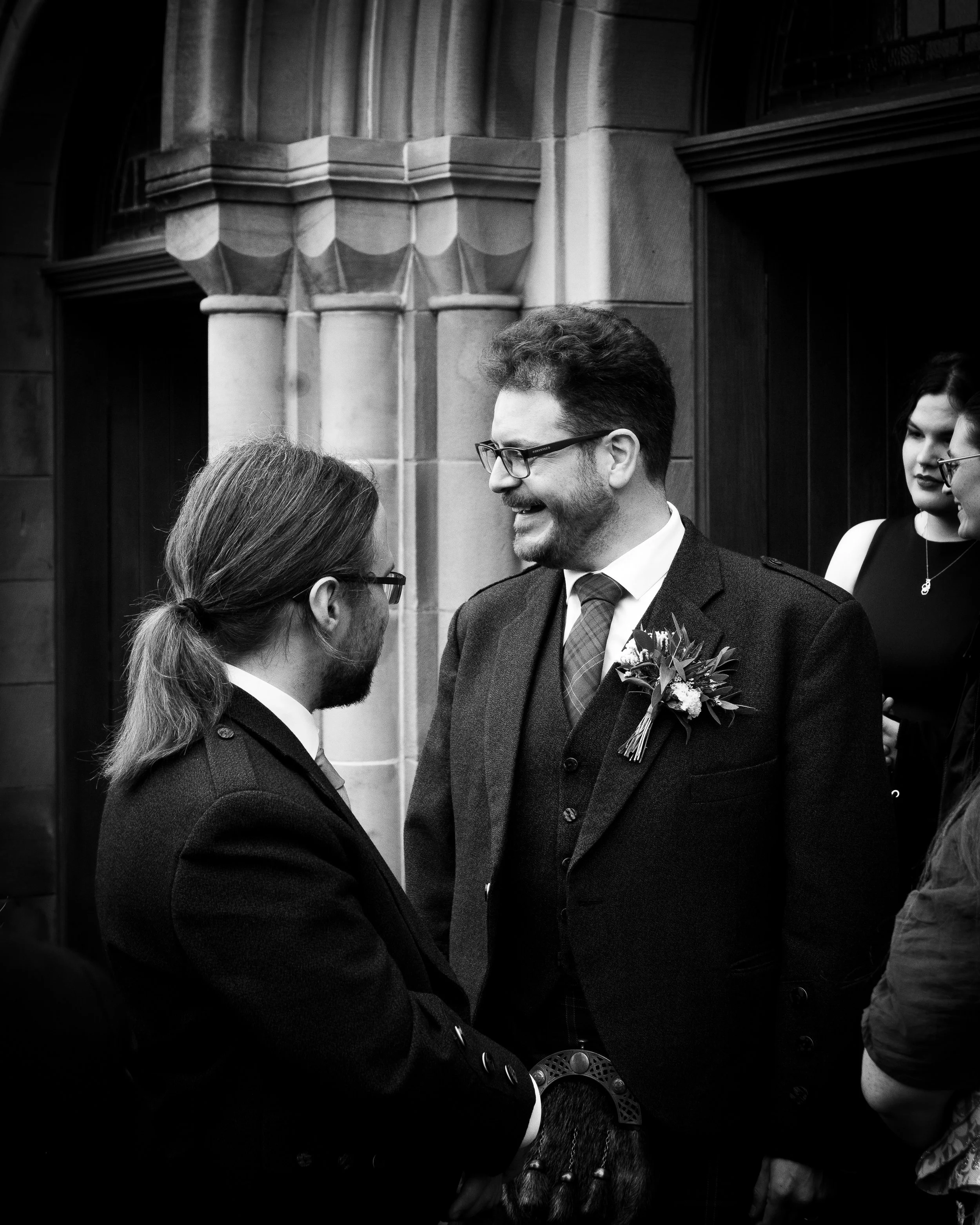 Two men at a wedding, smiling and talking to each other indoors with people in the background.