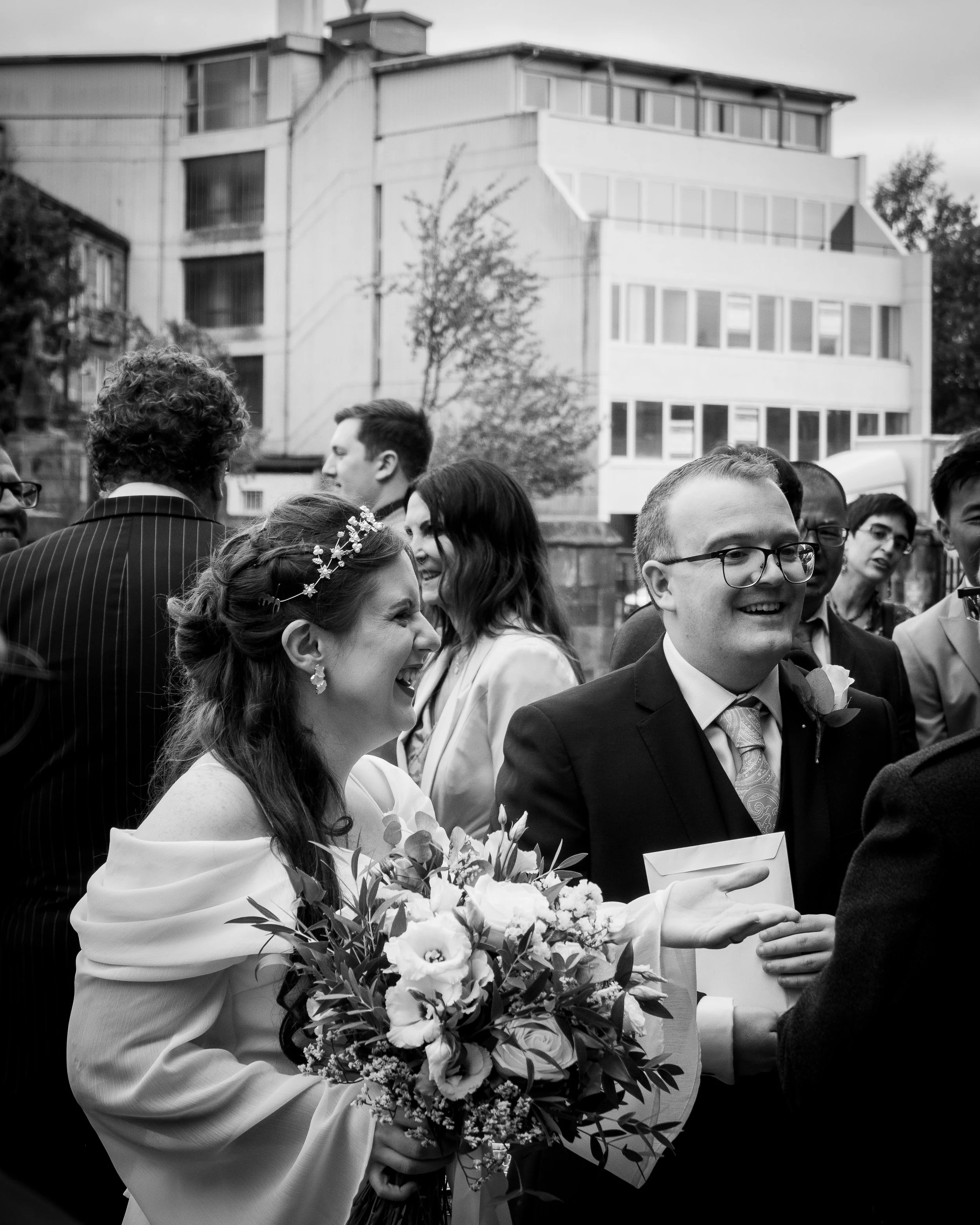 Black and white photo of a wedding celebration outdoors, showing a smiling bride holding a bouquet and engaging with a smiling groom wearing glasses and a suit.