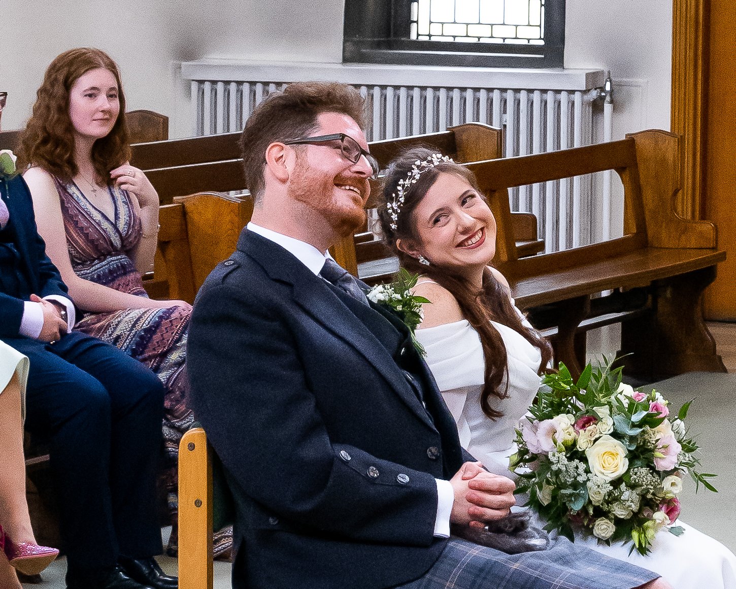 A bride and groom sitting happily during their wedding ceremony, with the bride holding a bouquet of flowers and smiling at the camera, and the groom leaning back and smiling. In the background, a woman with red hair and a patterned dress watches.