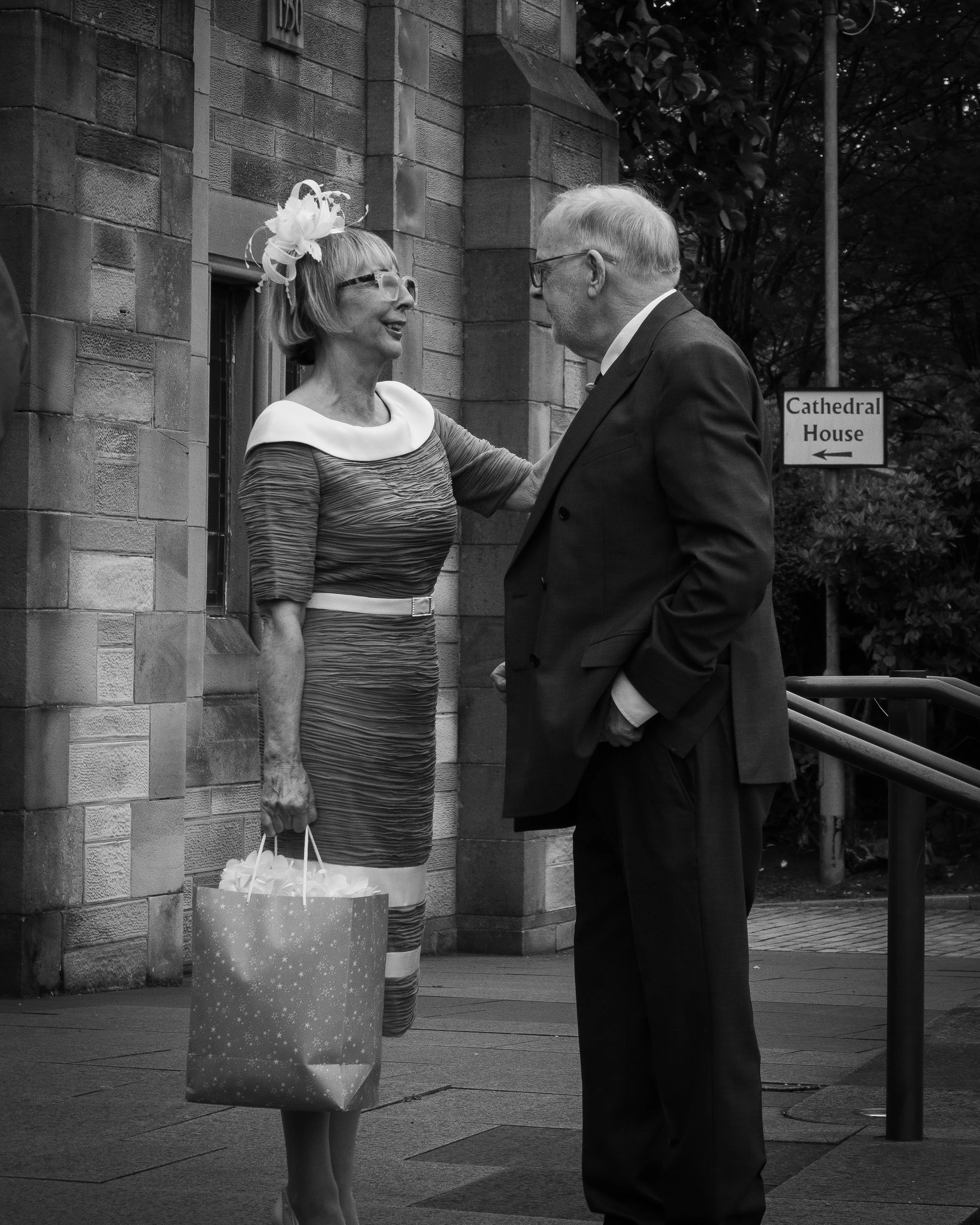 A woman in a dress and large fascinator hat holding a gift bag, smiling and speaking with an older man in a suit outside near a church marked "Cathedral House."