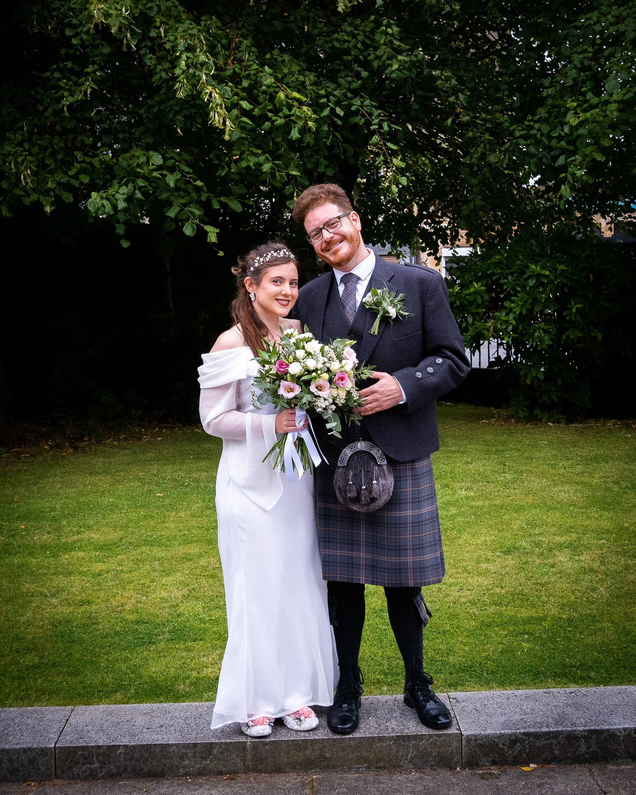 A bride and groom standing outdoors on a grassy area beneath a large leafy tree, smiling at the camera. The bride is holding a bouquet of flowers and is dressed in a white off-the-shoulder wedding gown. The groom is wearing a dark jacket, tie, and tr
