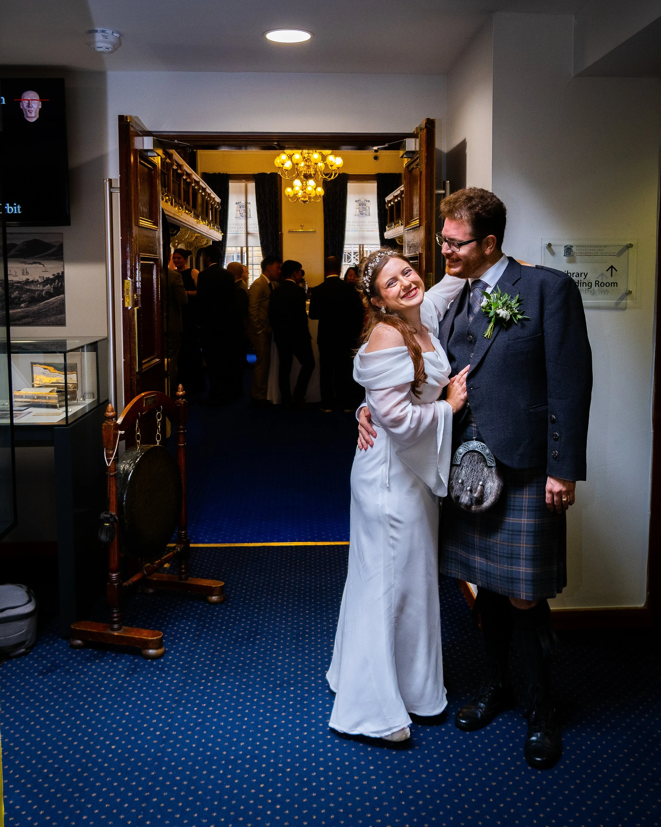 A woman in a white dress and a man in traditional Scottish attire are smiling and embracing at a wedding reception, with wedding guests in the background inside a decorated venue.