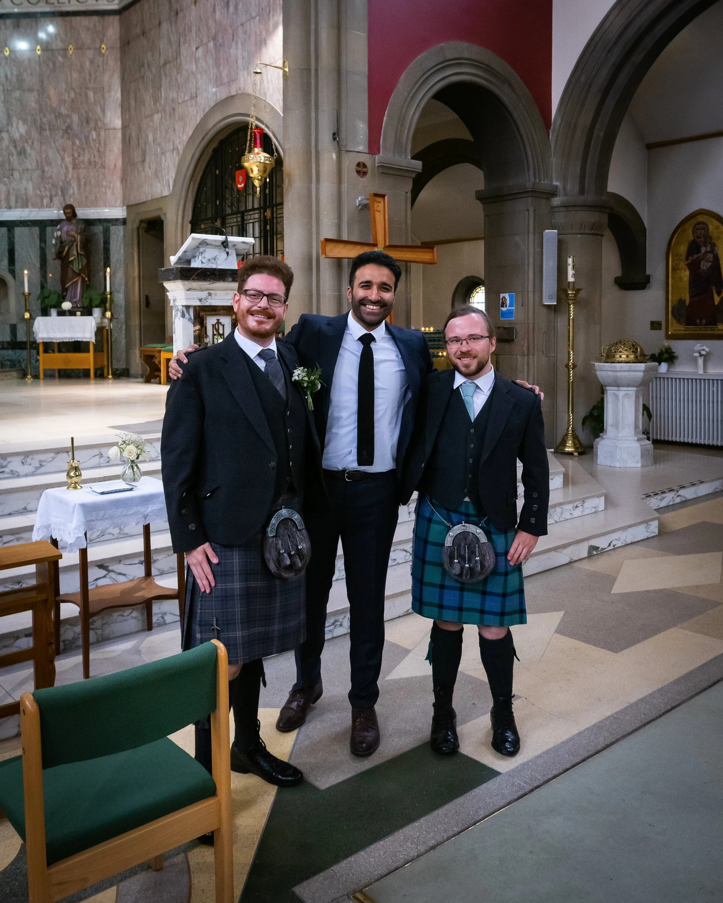 Three men dressed in suits, two wearing kilts, standing inside a church with an altar and religious symbols in the background.