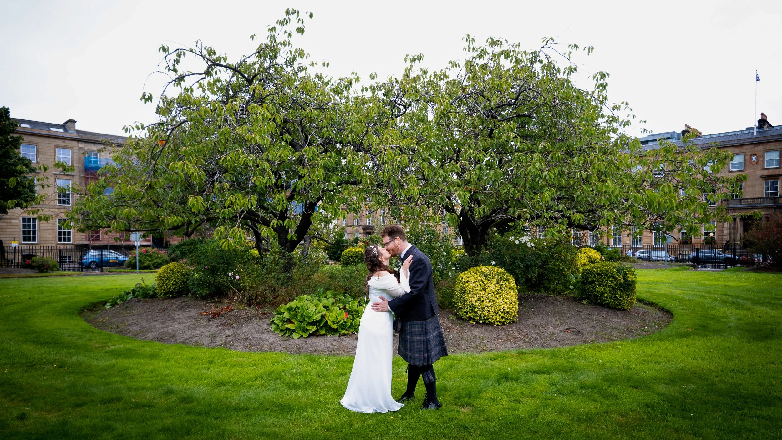 A couple dressed in wedding attire sharing a kiss in a garden with trees and shrubs, with residential buildings in the background.
