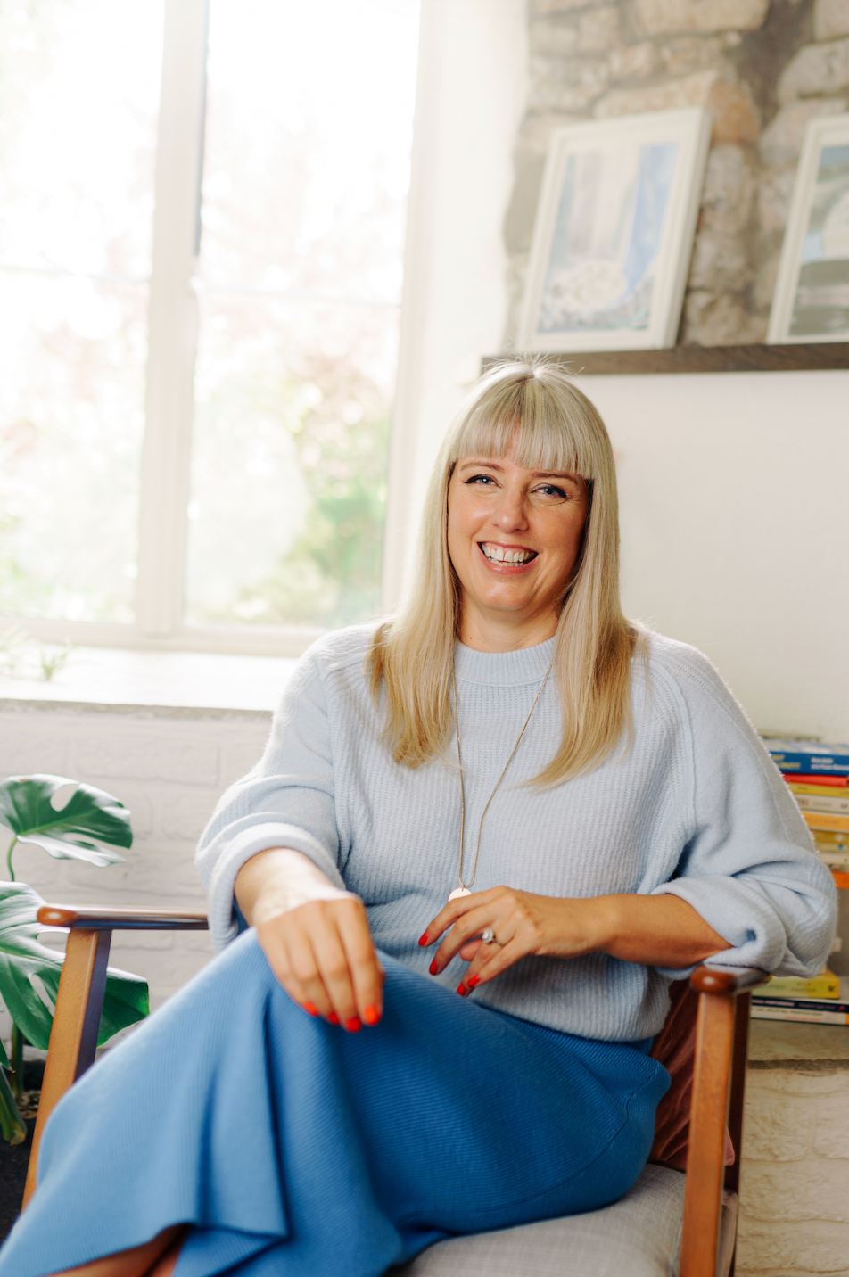 Caroline Allen Counselling - a woman with blonde hair smiling while sitting on a chair with a light-coloured cushion in a cozy room with large windows, framed pictures on the wall, a plant, and a stack of books.