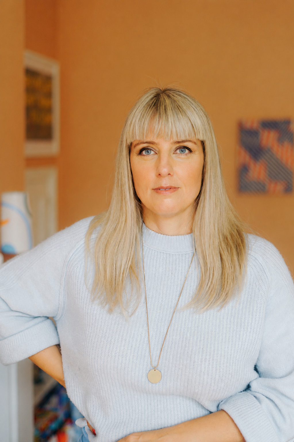 Caroline Allen Therapist- a woman with blonde hair and blue eyes wearing a white sweater and a long necklace, standing indoors, in front of a peach-coloured wall with artwork and a bookshelf visible in the background.