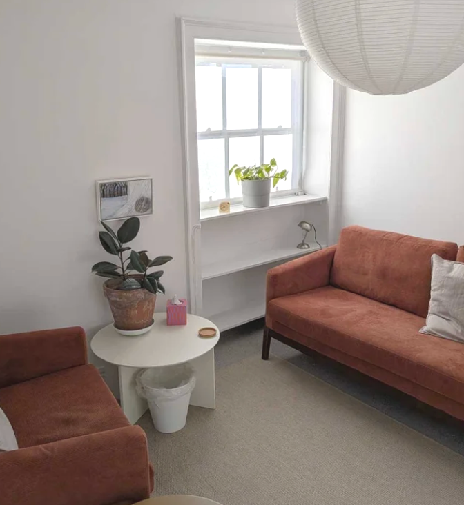 Caroline Allen's Therapy room on Park Street, Bristol, with two terracotta couches, a white side table with a potted plant, and a window with a white sill and potted plant.