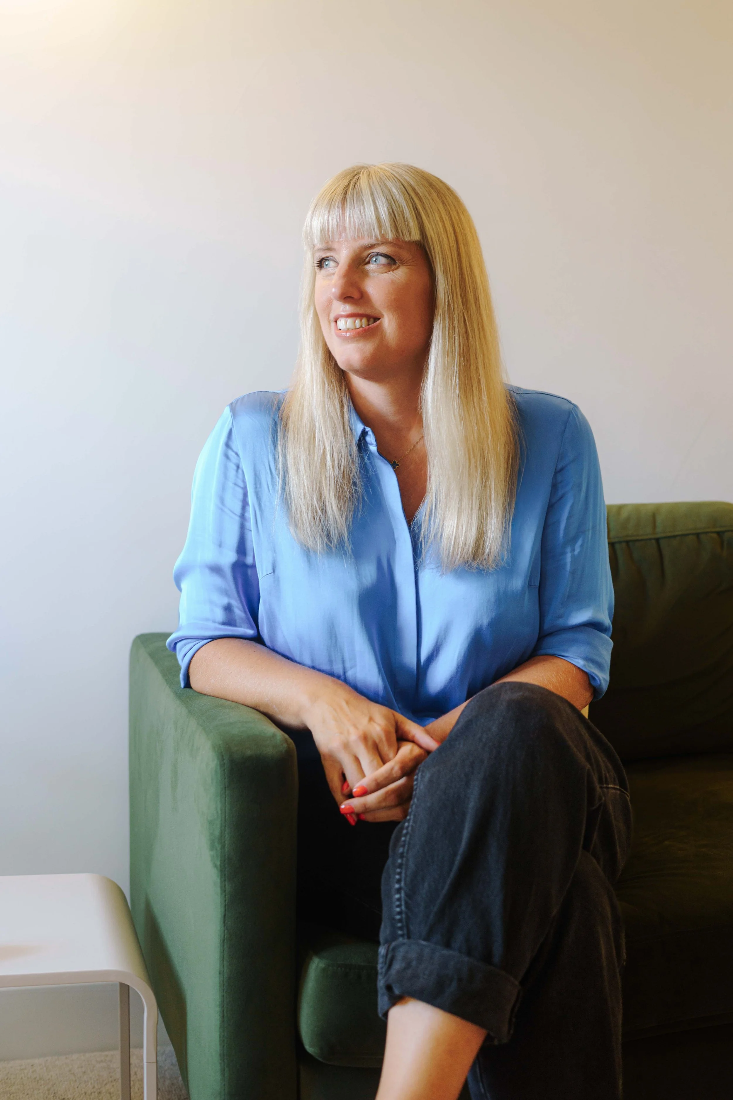 Caroline Allen-a woman with long blonde hair and wearing a blue blouse, sitting on a green couch in her Therapy room on Park Street, Bristol, smiling and looking to the side.