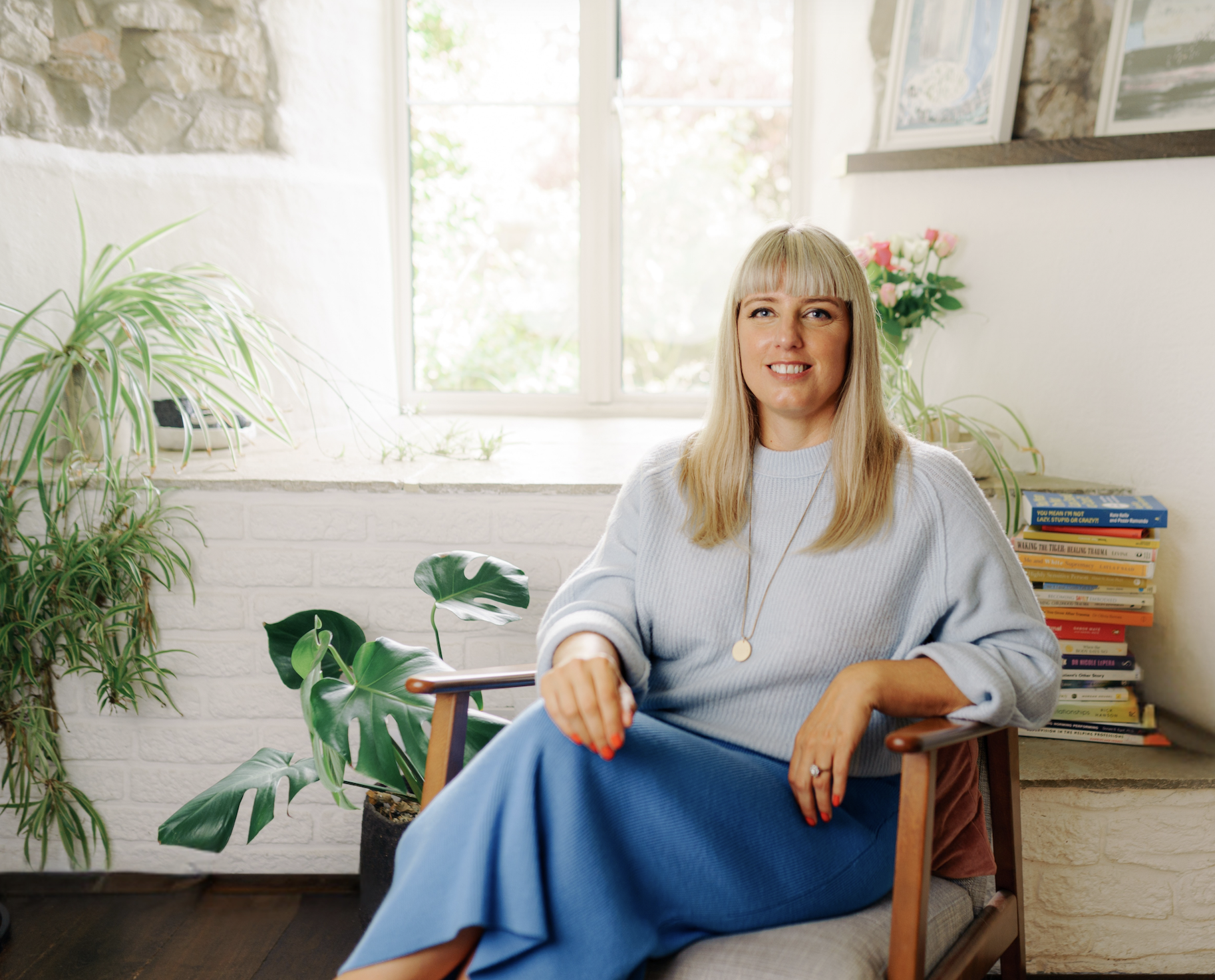 Caroline Allen Therapeutic Counselling -  a woman with blonde hair, smiling while sitting in a cozy indoor space, surrounded by plants and books on a shelf.