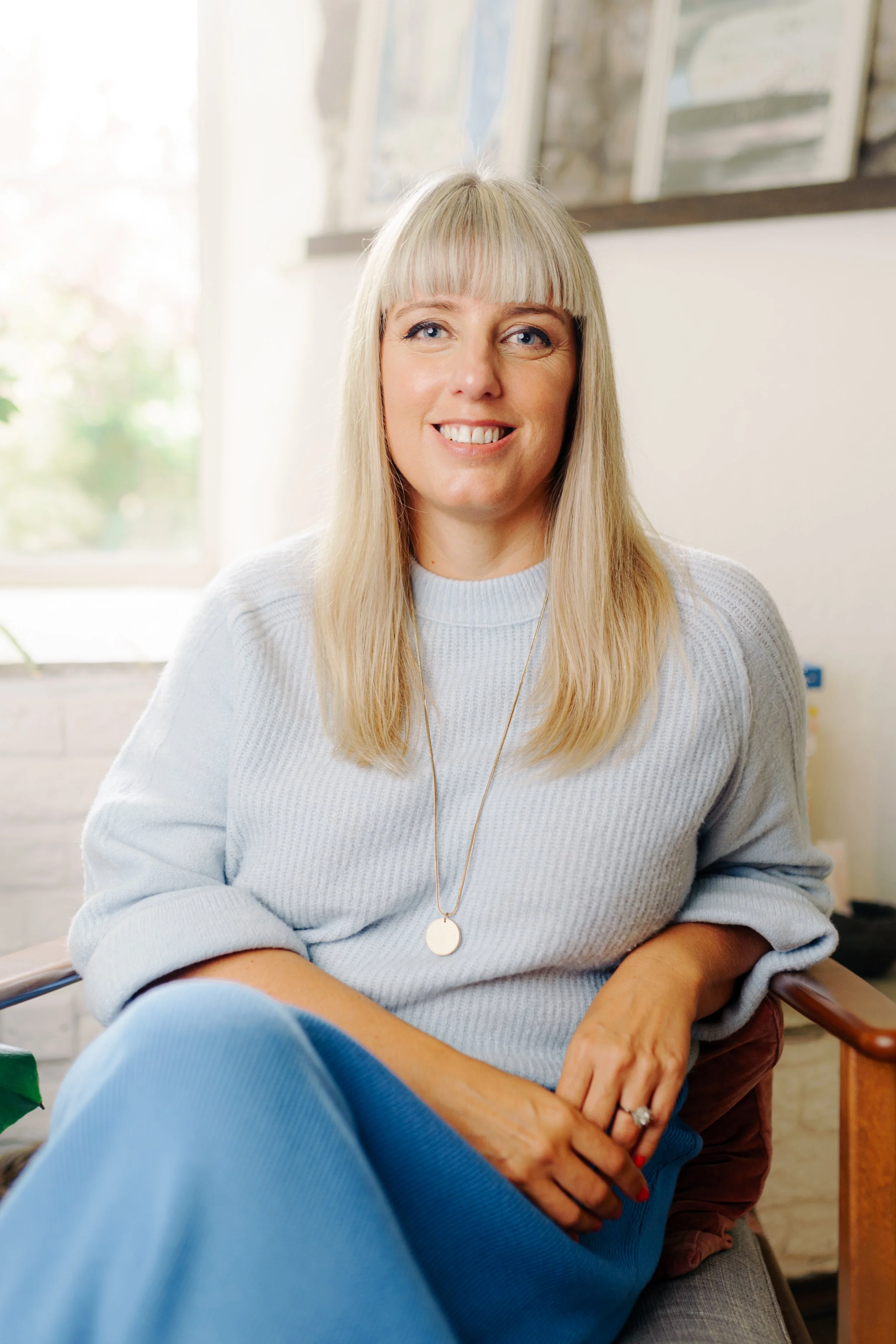 Caroline Allen Counselling - a woman with long blonde hair, wearing a light grey sweater and a round pendant necklace, is sitting in a well-lit room with large windows. She is smiling and looking at the camera.