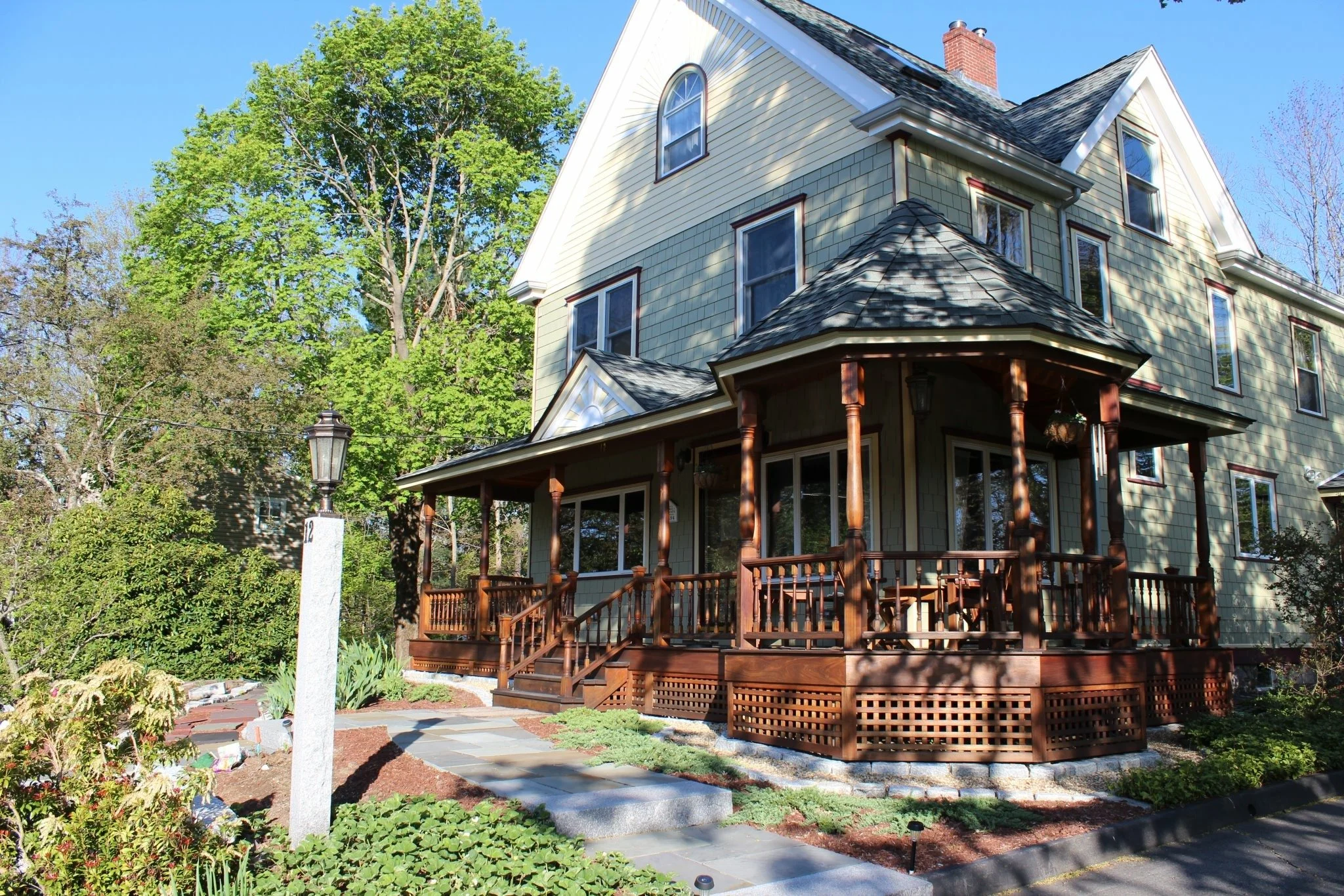 A multi-story house with a large front porch, surrounded by a lush green garden and tall trees, under a clear blue sky.