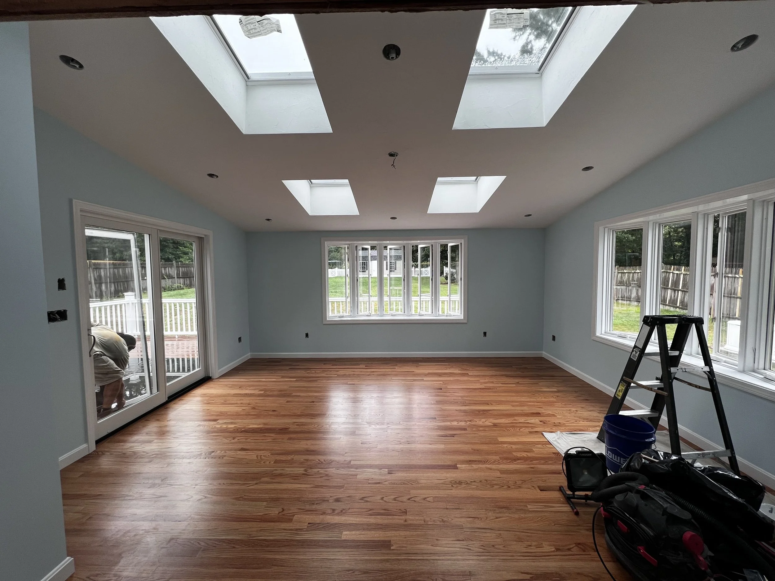 Empty living room with light blue walls, hardwood floor, multiple large windows, and skylights in the ceiling. Construction tools and equipment are visible on the right side of the room.