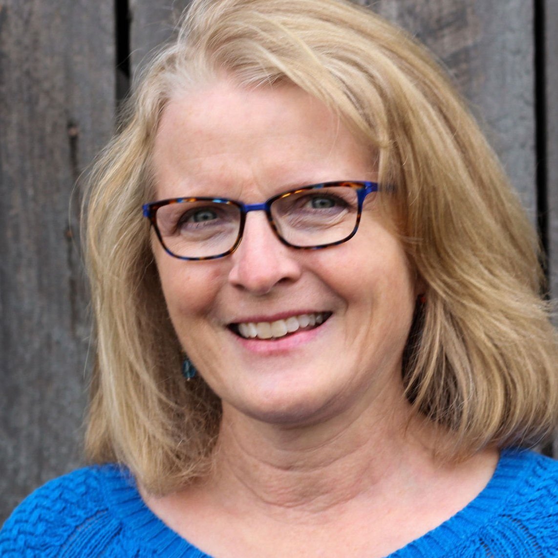 Close-up of a smiling woman with blonde hair, wearing glasses with tortoiseshell and blue frames, a blue top, standing in front of a wooden fence.