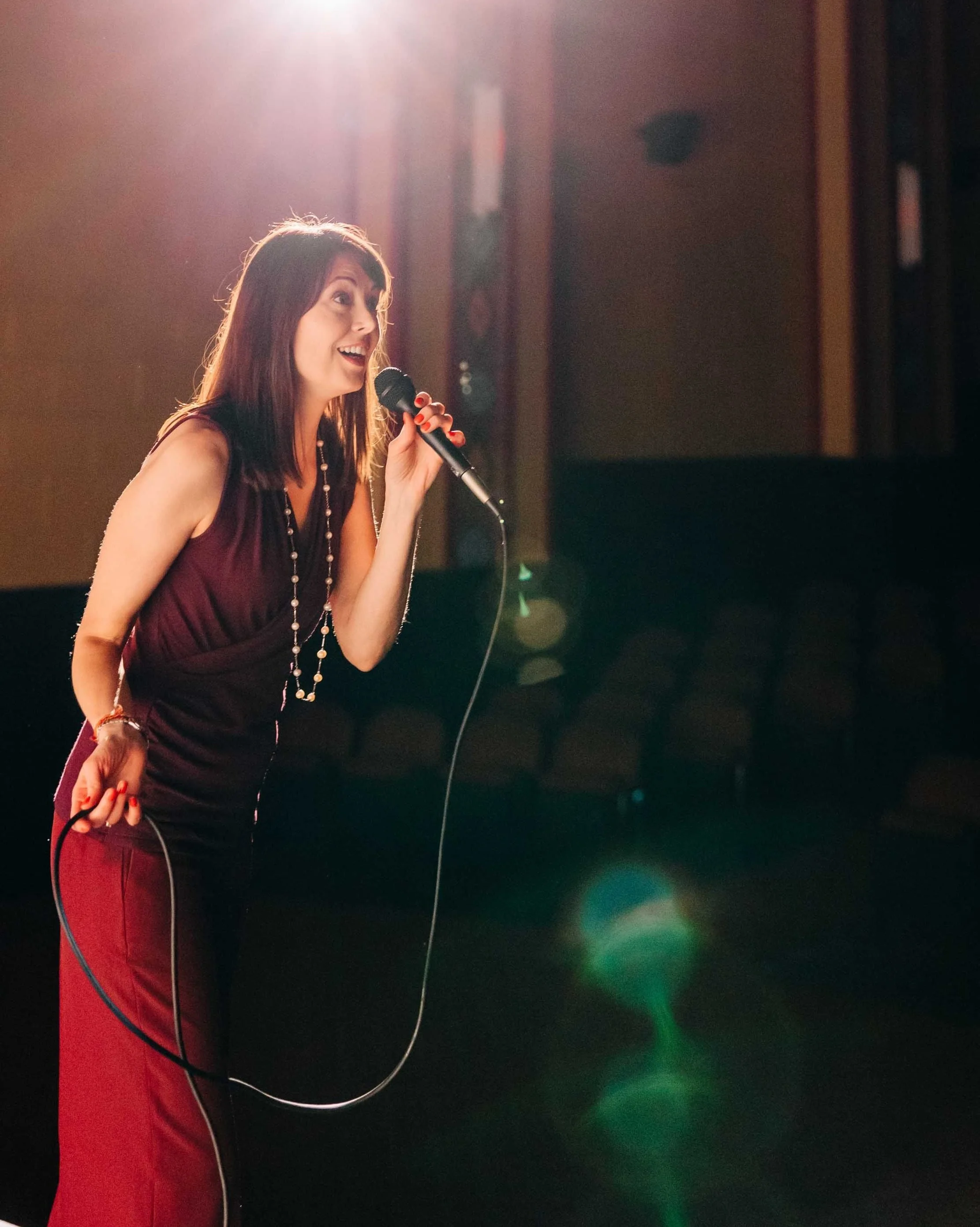 A woman singing into a microphone on stage, illuminated by stage lights with an empty audience area in the background.