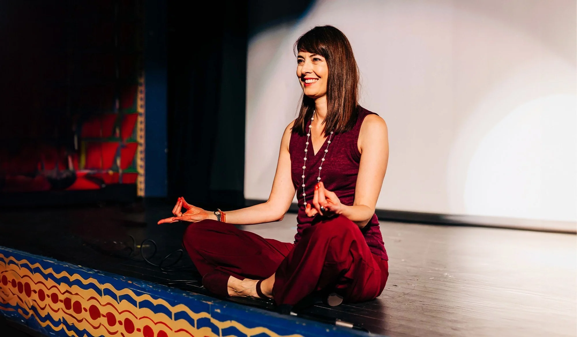 A woman with dark brown hair, sitting cross-legged on stage, wearing a sleeveless maroon top and matching maroon pants, smiling and meditating with a beaded necklace and bracelets, on an empty stage with a colorful painted border.