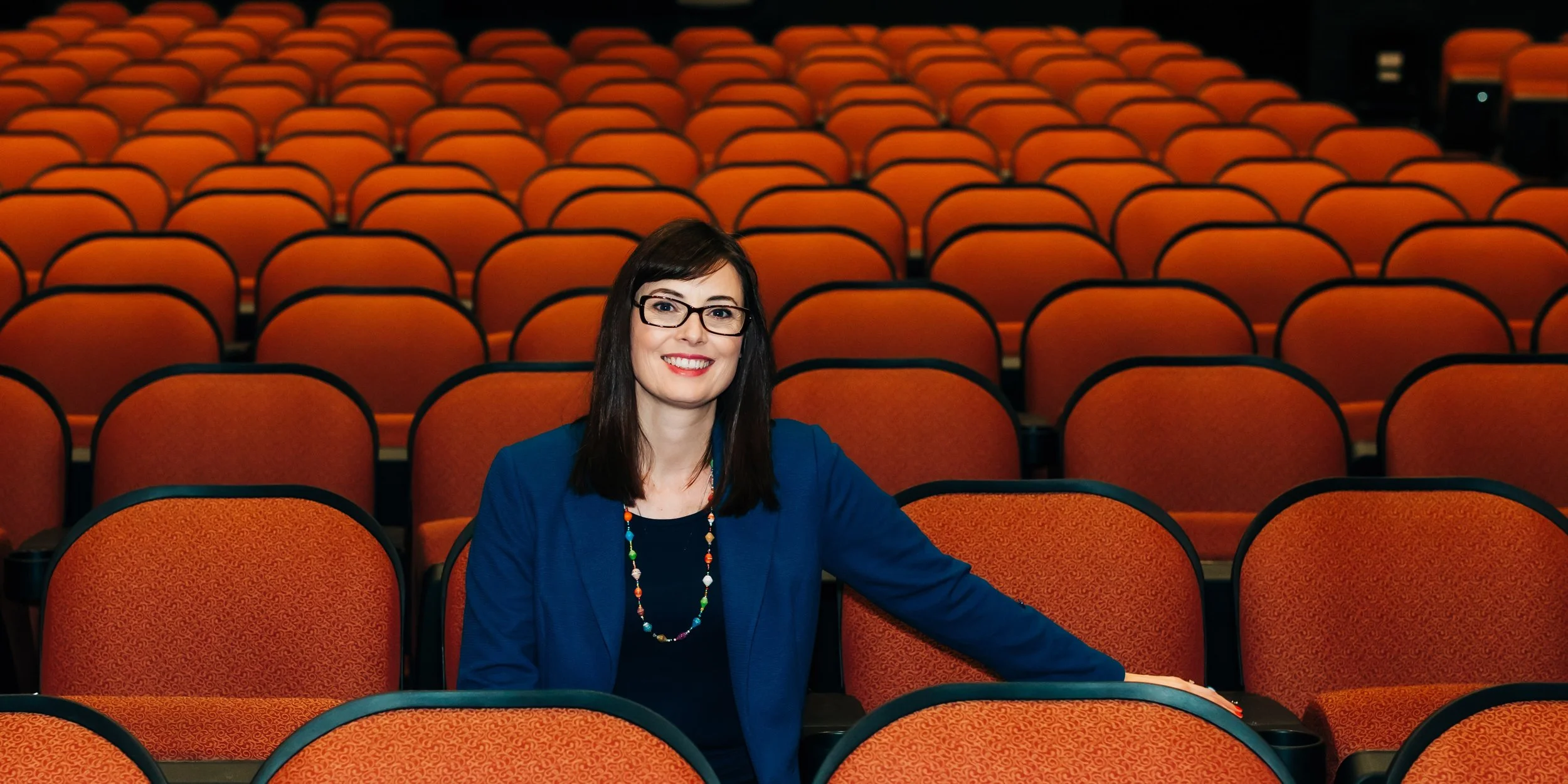 A woman with shoulder-length dark brown hair, smiling, wearing a sleeveless maroon top and a pearl necklace, standing against a white and black background.