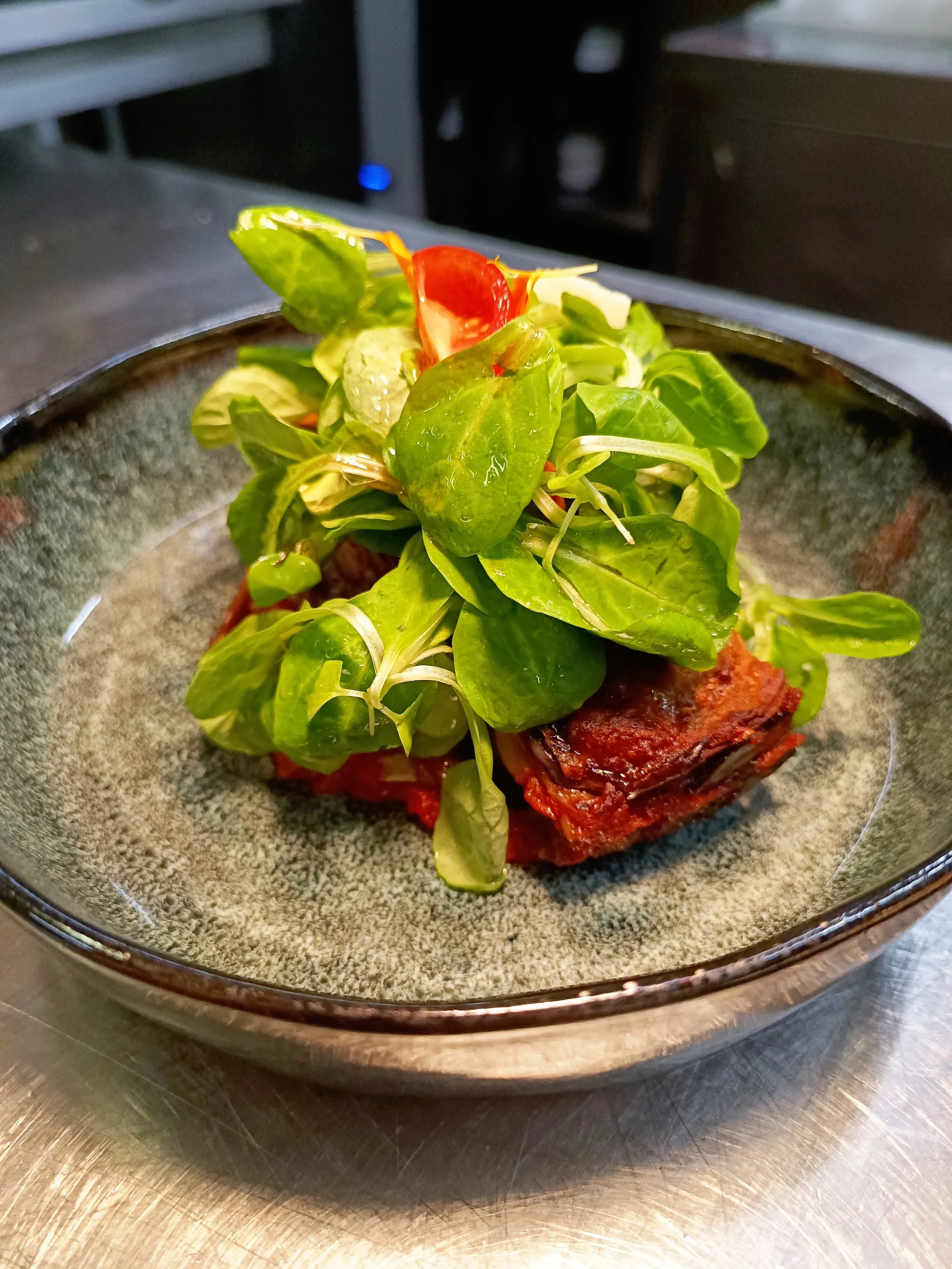 Un plat composé de jeunes pousses de salade sur une couche de légumes cuits, des tomates et des aubergines, dans une assiette gris foncé.