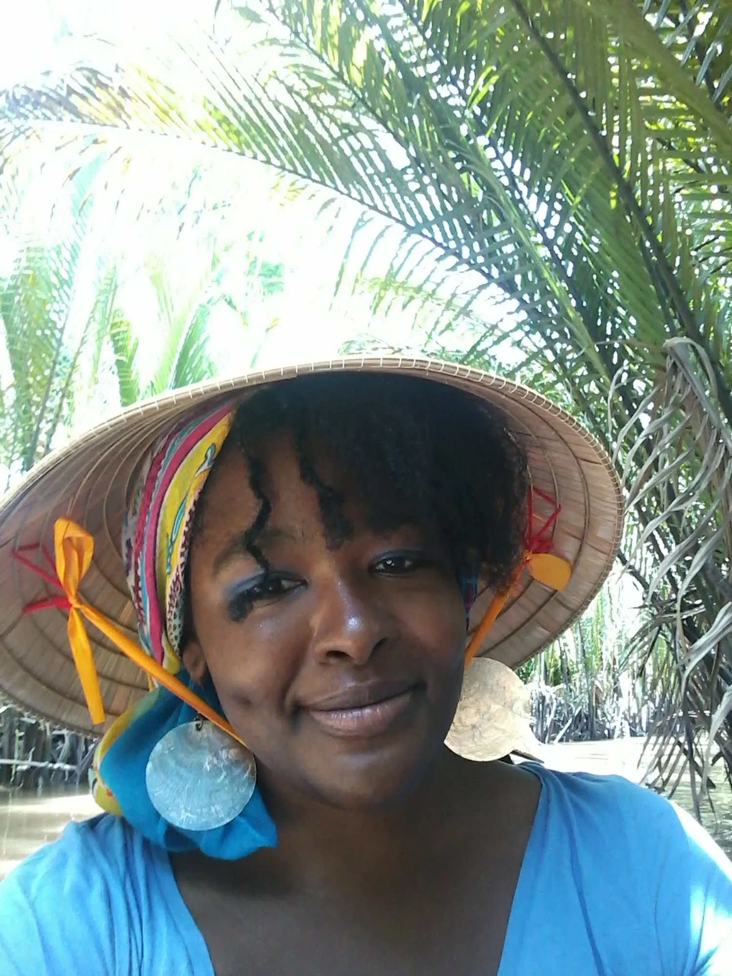 Black woman wearing a large straw hat with colorful accents and big circular earrings, standing among lush green palm trees.