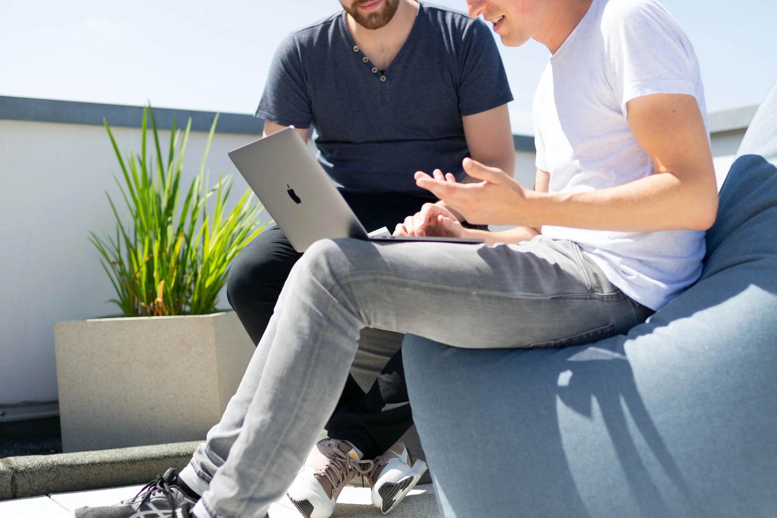 Two men sitting outdoors, one holding a laptop, having a discussion next to a plant.