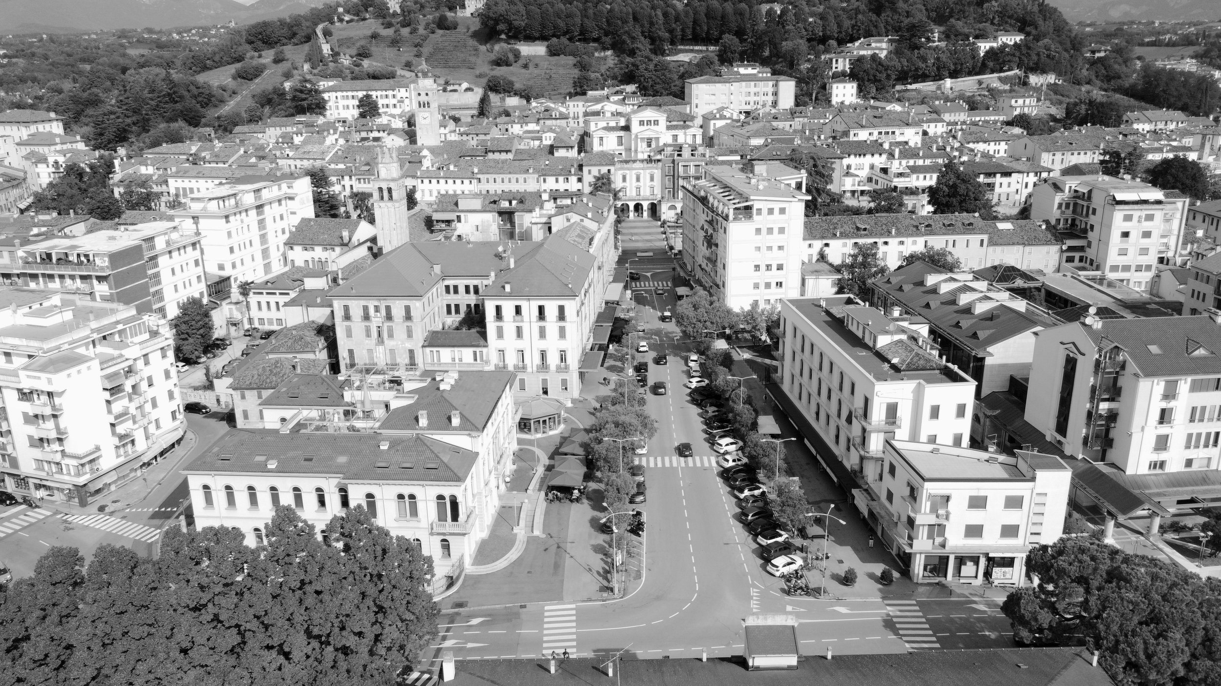 Veduta aerea di una città con edifici residenziali e commerciali, strade con auto parcheggiate e alberi lungo le vie, colline sullo sfondo.