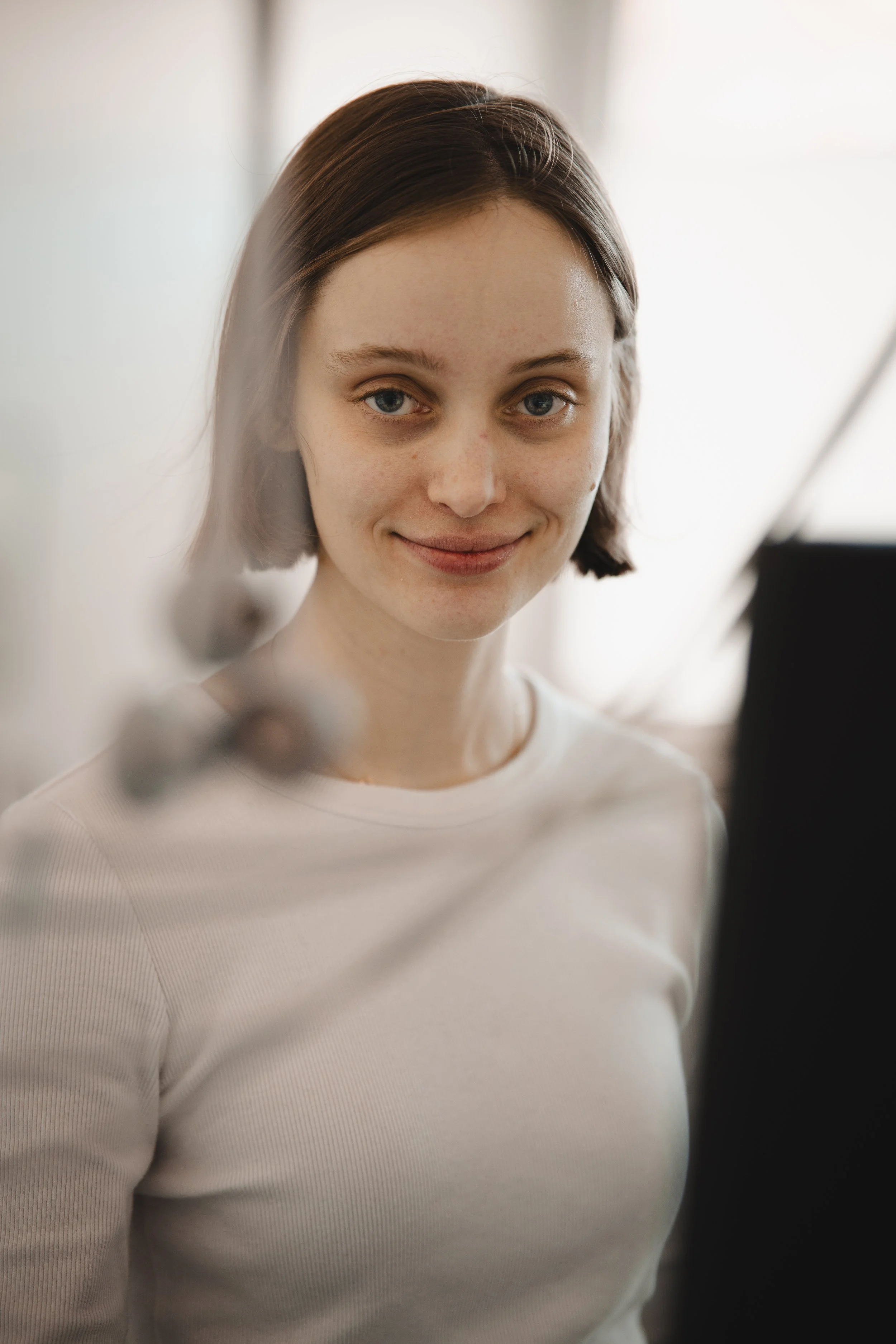 Junge Frau mit braunen Haaren und hellem Hemd steht vor einer transparenten Oberfläche und lächelt. Blickrichtung direkt in die Kamera.