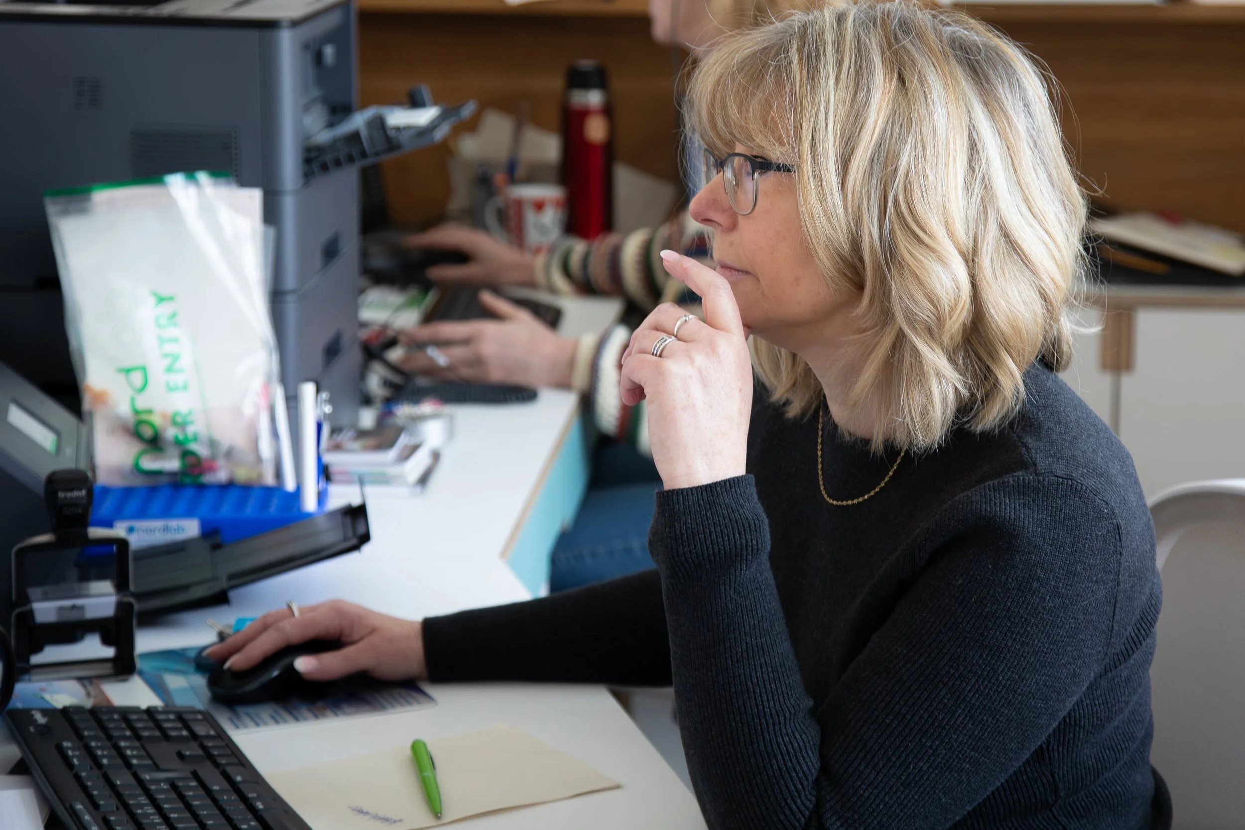 Frau mit Brille und blonden Haaren arbeitet an einem Computer in einem Büro, neben ihr eine Packung Popcorn.