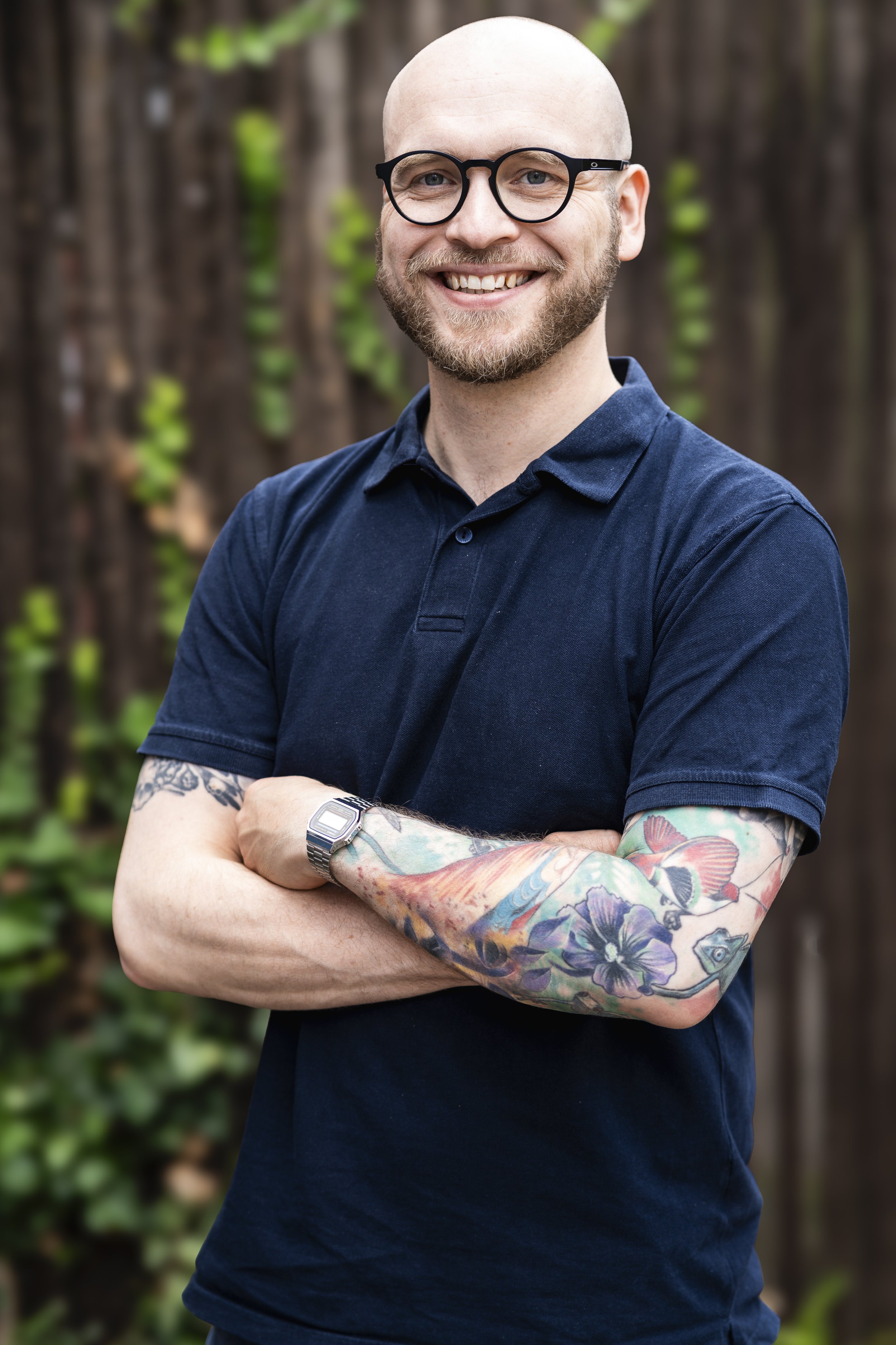 Ein lächelnder Mann mit Brille, Tattoos am Arm, trägt eine Armbanduhr und ein dunkelblaues T-Shirt, steht vor einem Holzzaun mit grünen Pflanzen im Hintergrund.