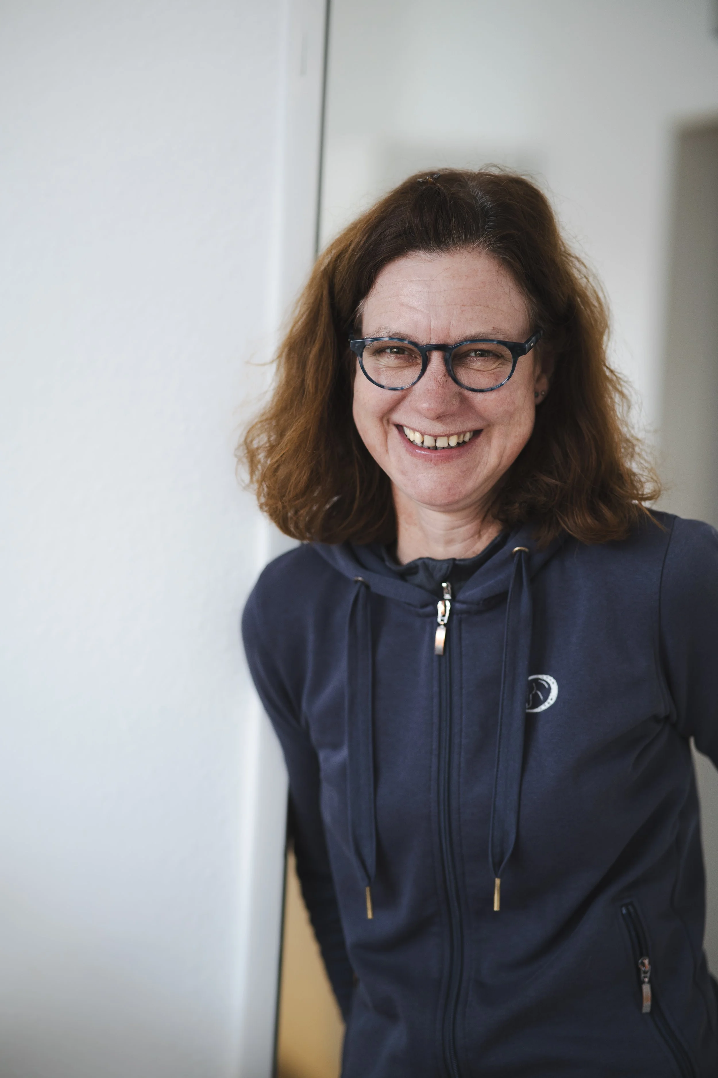 Freudestrahlende Frau mit Brille in dunkelblauem Hoodie, lächelnd, steht neben einer weißen Wand.
