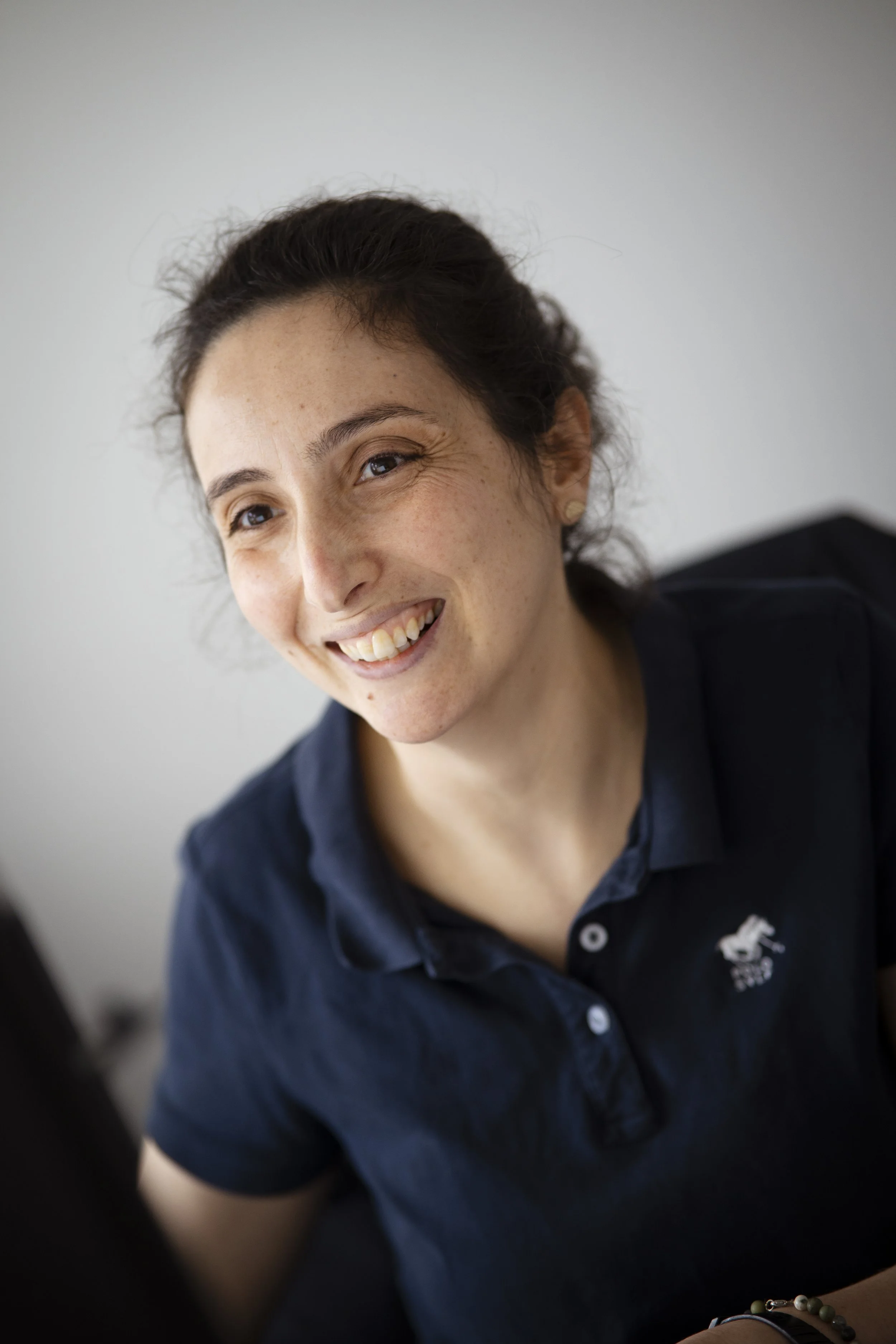 Nahaufnahme einer lachenden jungen Frau mit dunklen Haaren und Sommersprossen, die einen dunklen Polo trägt.