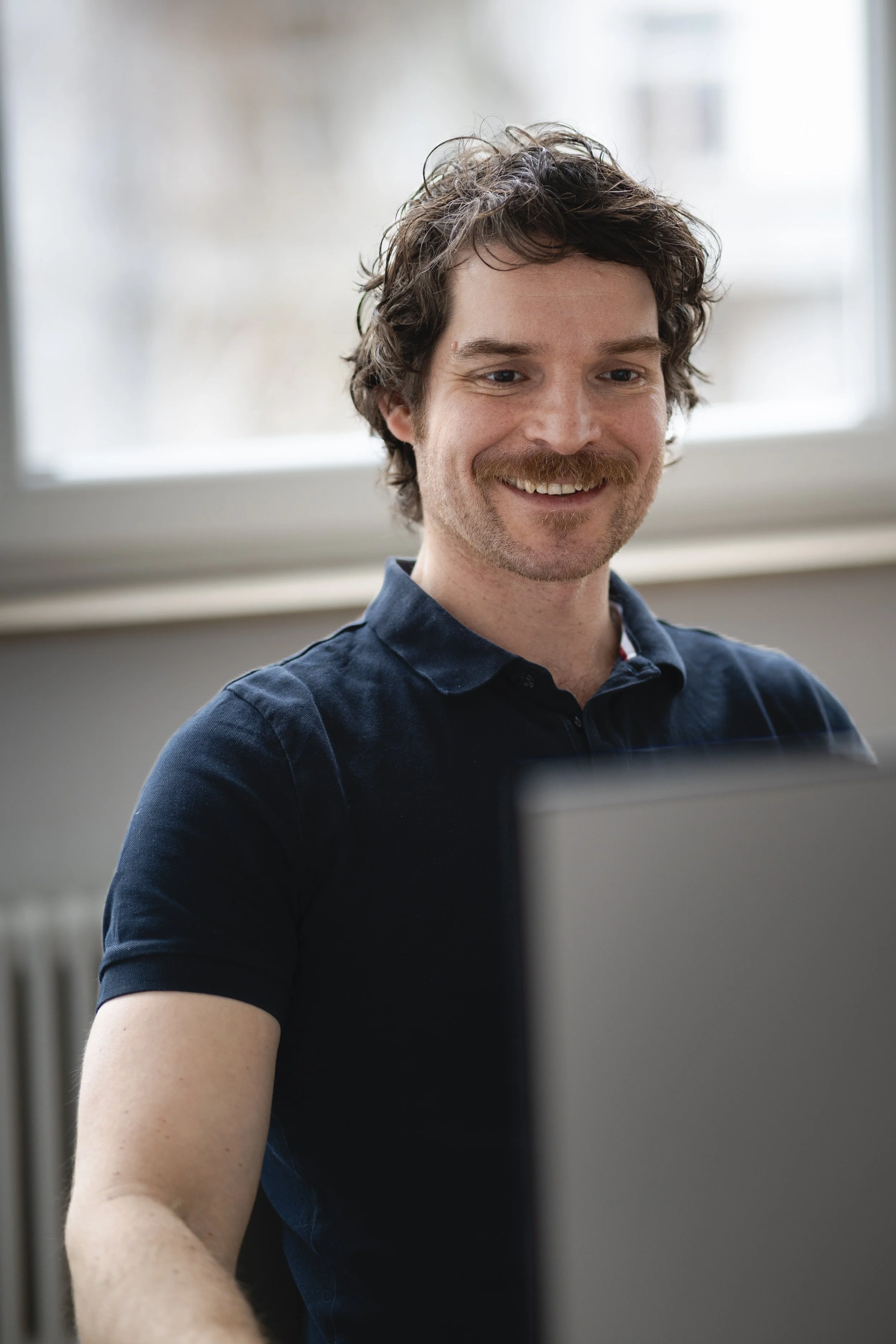 Mann mit dunklem Hemd sitzt vor einem Computer und lächelt.