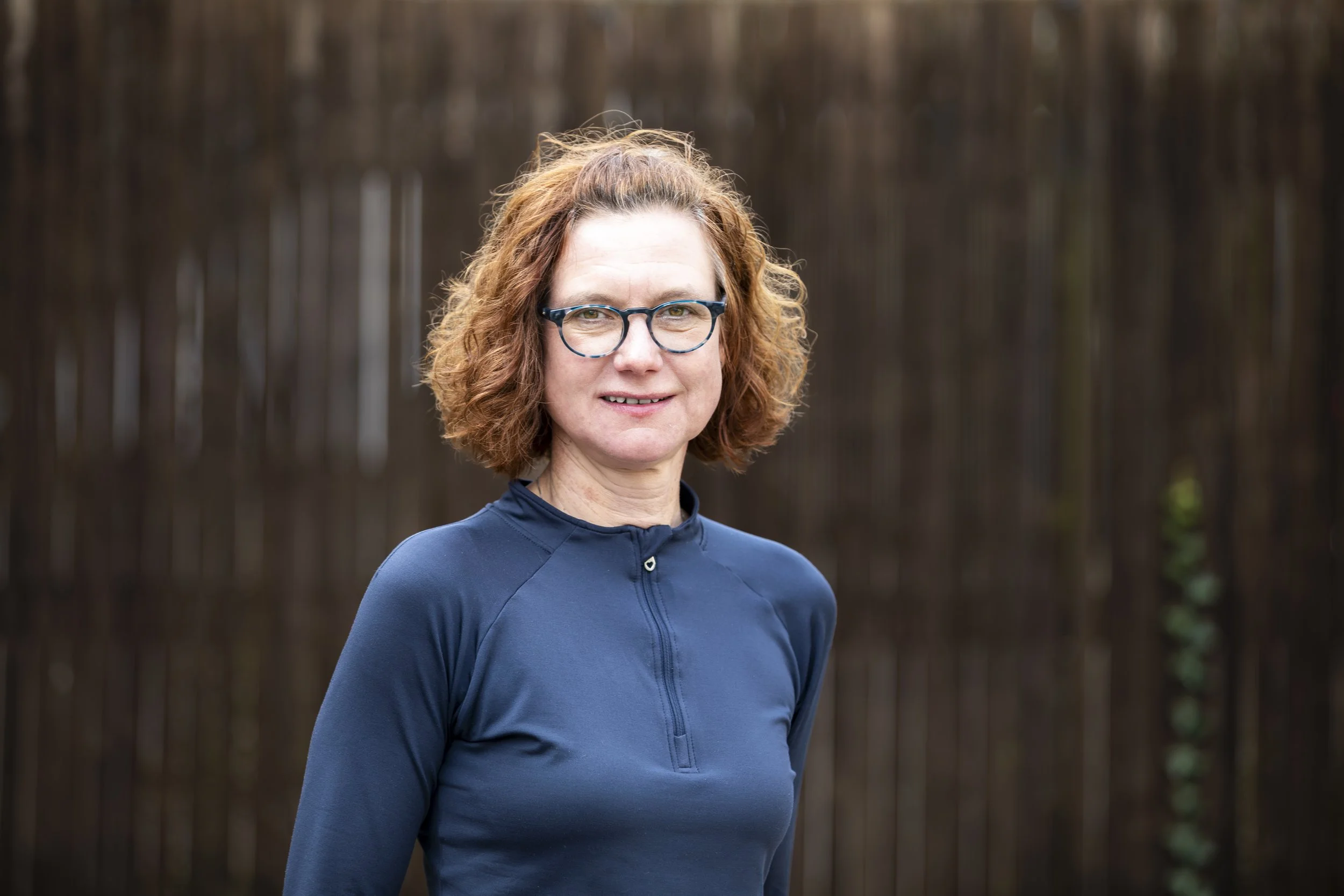 Frau mit roten, lockigen Haaren, Brille und blauer Sportjacke steht vor einem Holzzaun.