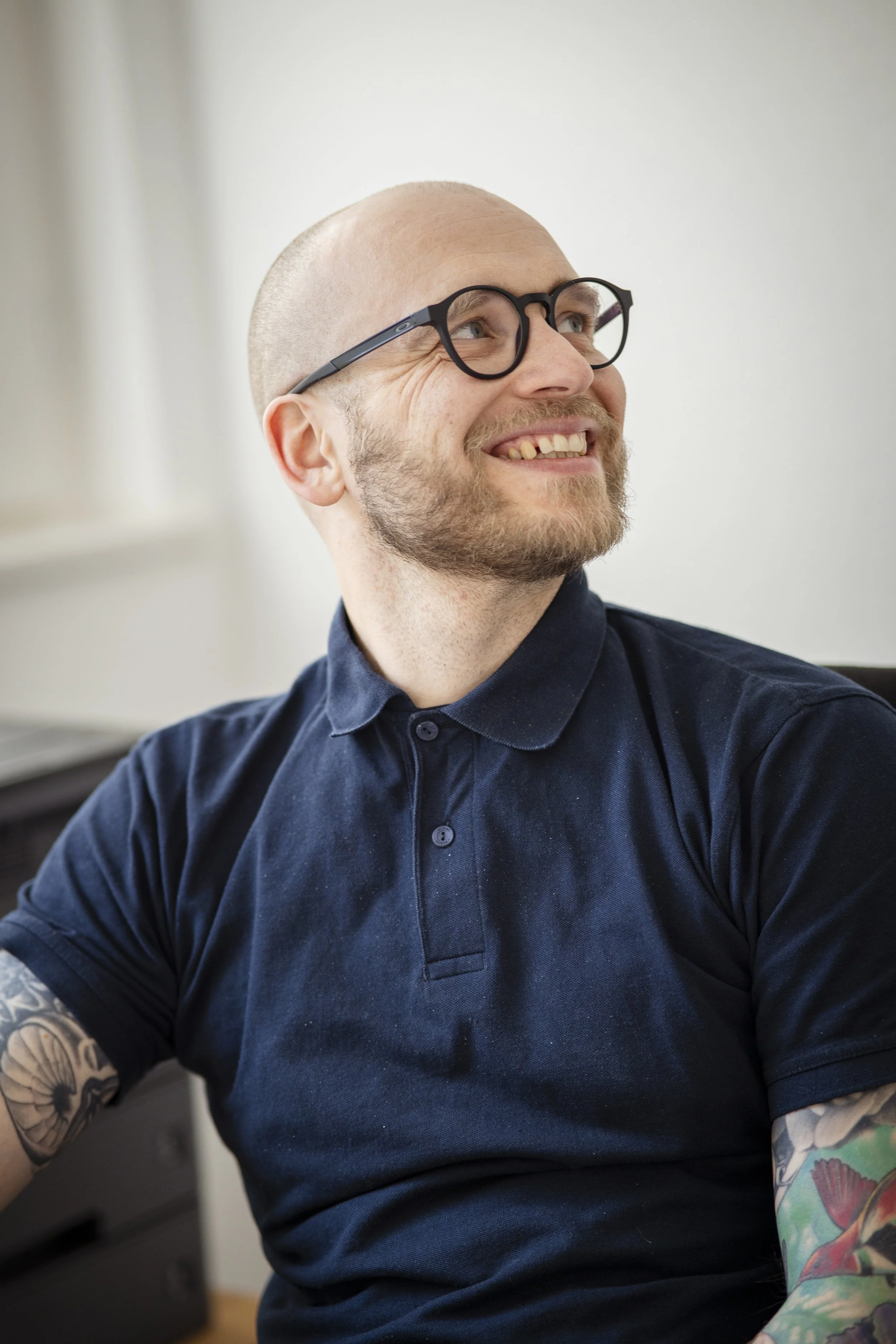 Lächelnder Mann mit Brille, Glatzenkopf, Tätowierungen am Arm, trägt ein dunkelblaues Polo-Shirt, im Innenraum.