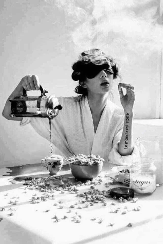 Une femme en peignoir verse du café dans une tasse, entourée de céréales, avec un bol de céréales, un sucre en poudre, et une cuillère sur une table. Elle porte des lunettes de soleil et a des cheveux ondulés.