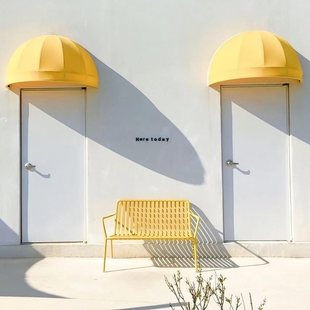 Deux portes blanches avec des auvents jaunes au-dessus, une banc jaune en dessous, et la phrase "Here today" écrite sur le mur.