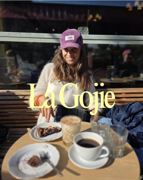 Une femme souriante portant une casquette violette, assise à une table de café avec des boissons et des desserts, dans un cadre extérieur.
