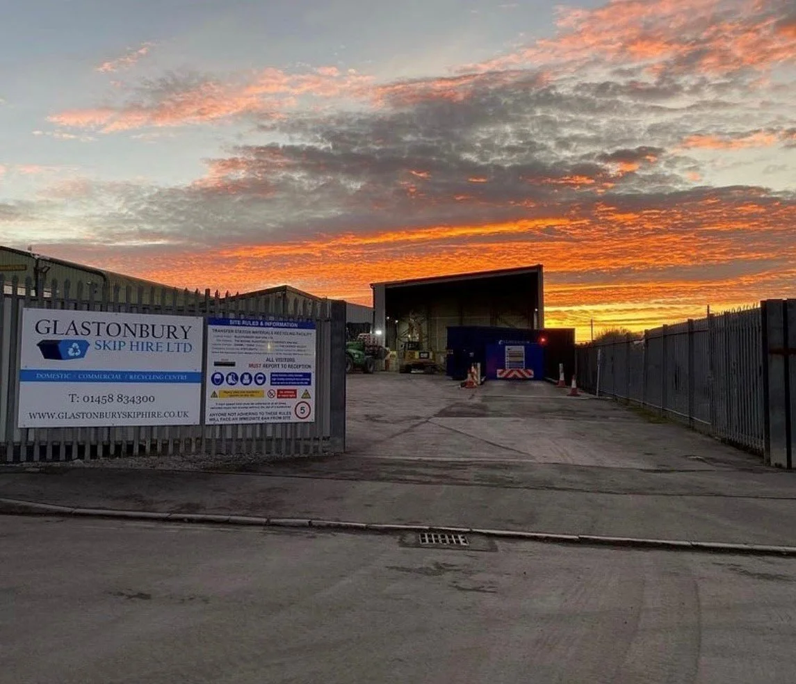 The image depicts an industrial area at sunset, with a vibrant orange and pink sky. There is a large open warehouse with some equipment inside, surrounded by a fence. A sign on the fence reads 'Glastonbury Skip Hire Ltd,' and the entrance is open with a paved driveway leading into the yard.