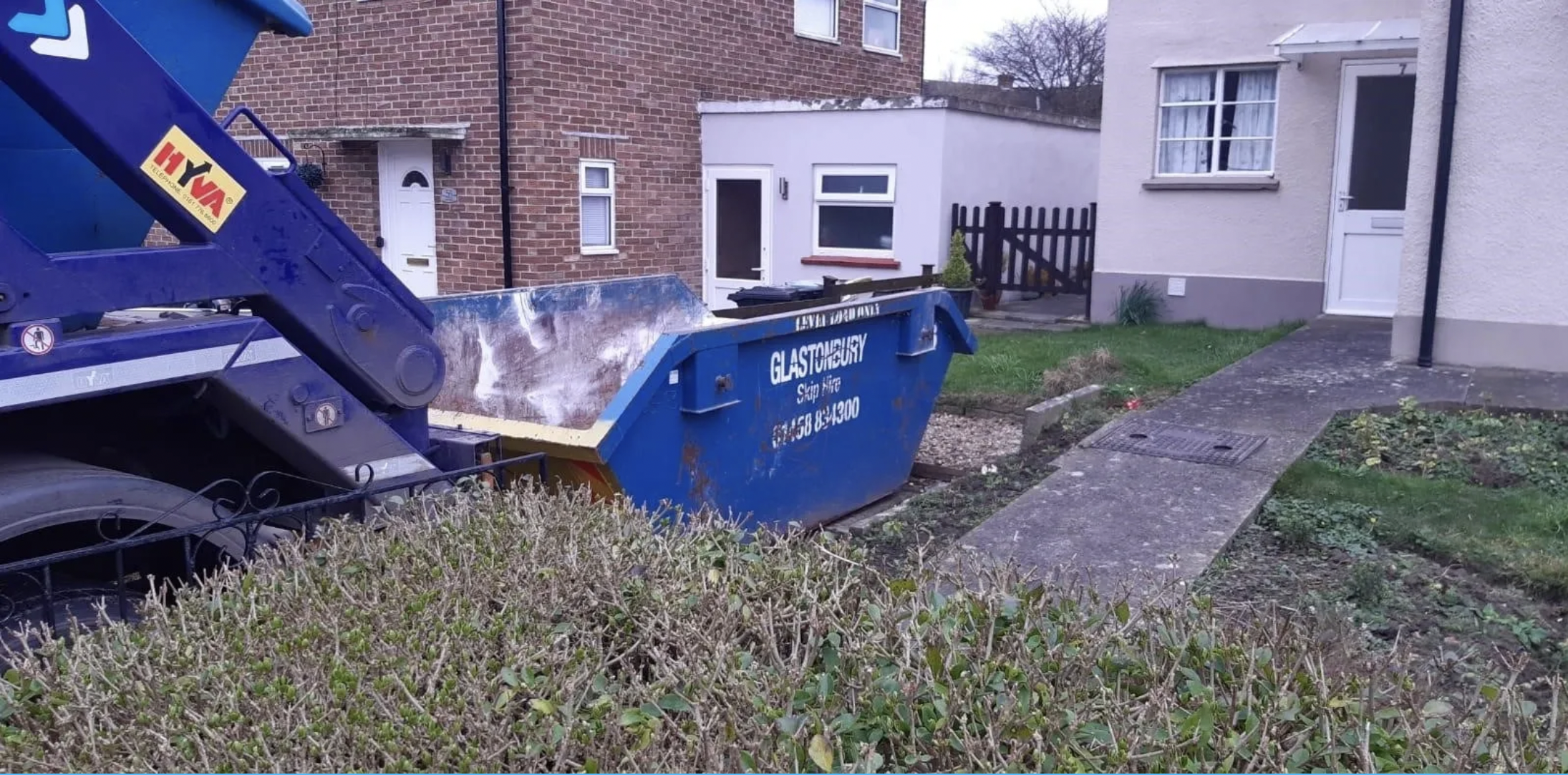 A blue dumpster outside residential houses, with a hedge in the foreground and brick and white houses in the background, during daytime.