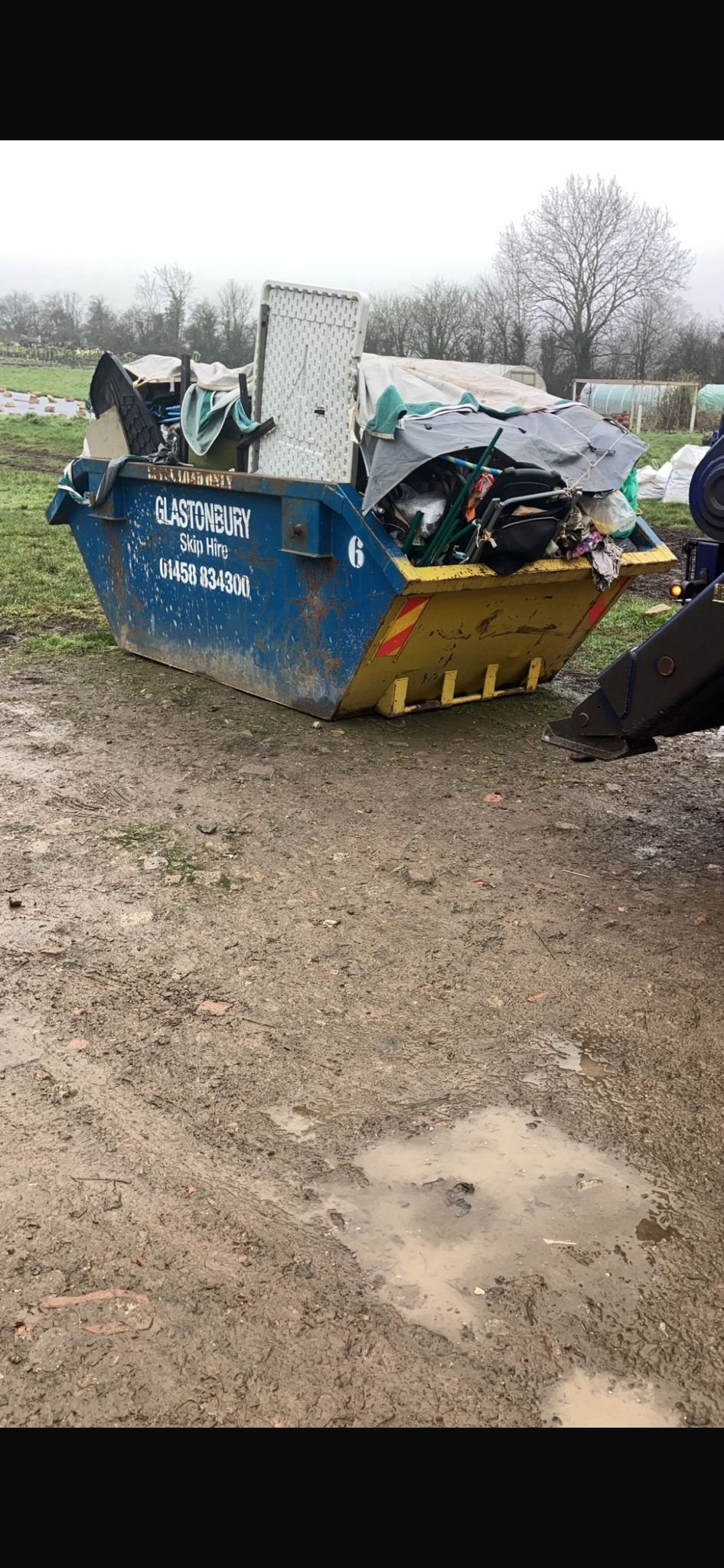 Blue skip bin filled with discarded furniture and debris, situated outdoors on muddy ground with puddles, in a rural area with trees and fields in the background.
